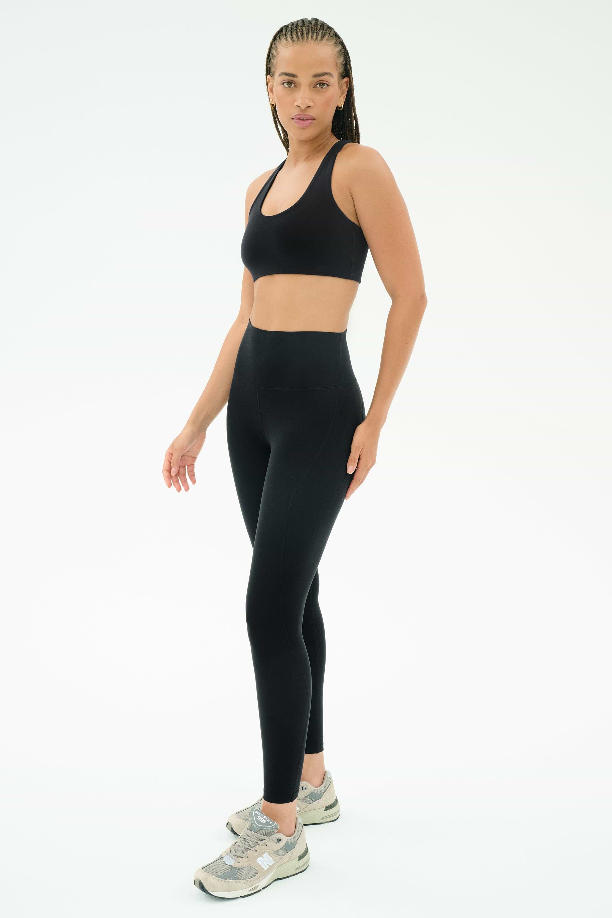 A woman wears the SPLITS59 Airweight High Waist 7/8 Pocket Legging in black, a matching sports bra, and gray sneakers, standing against a plain white background.