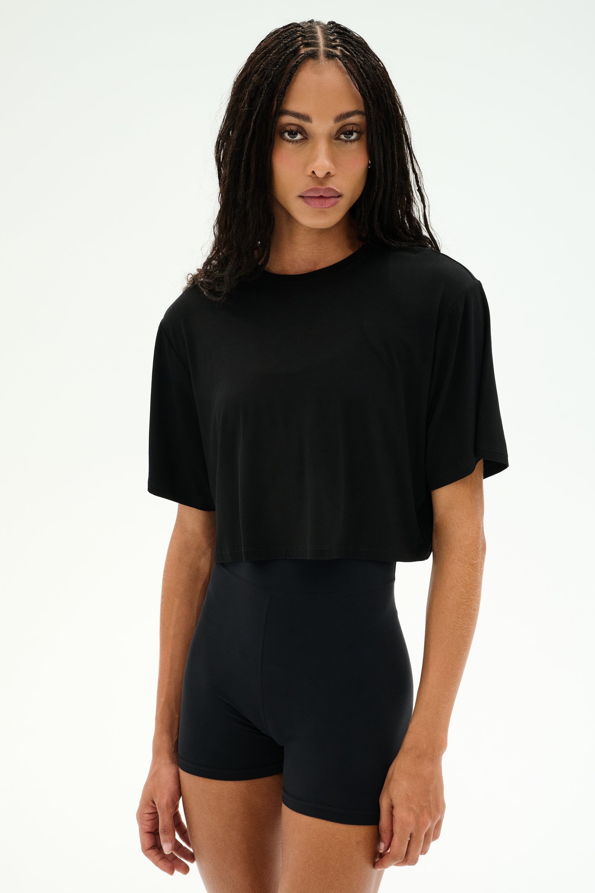 A person with long, braided hair wears the SPLITS59 Djuna Oversized Crop Jersey Tee in black and stretchy black high-waisted shorts, standing against a plain light background.