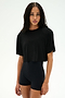 A person with long, braided hair wears the SPLITS59 Djuna Oversized Crop Jersey Tee in black and stretchy black high-waisted shorts, standing against a plain light background.