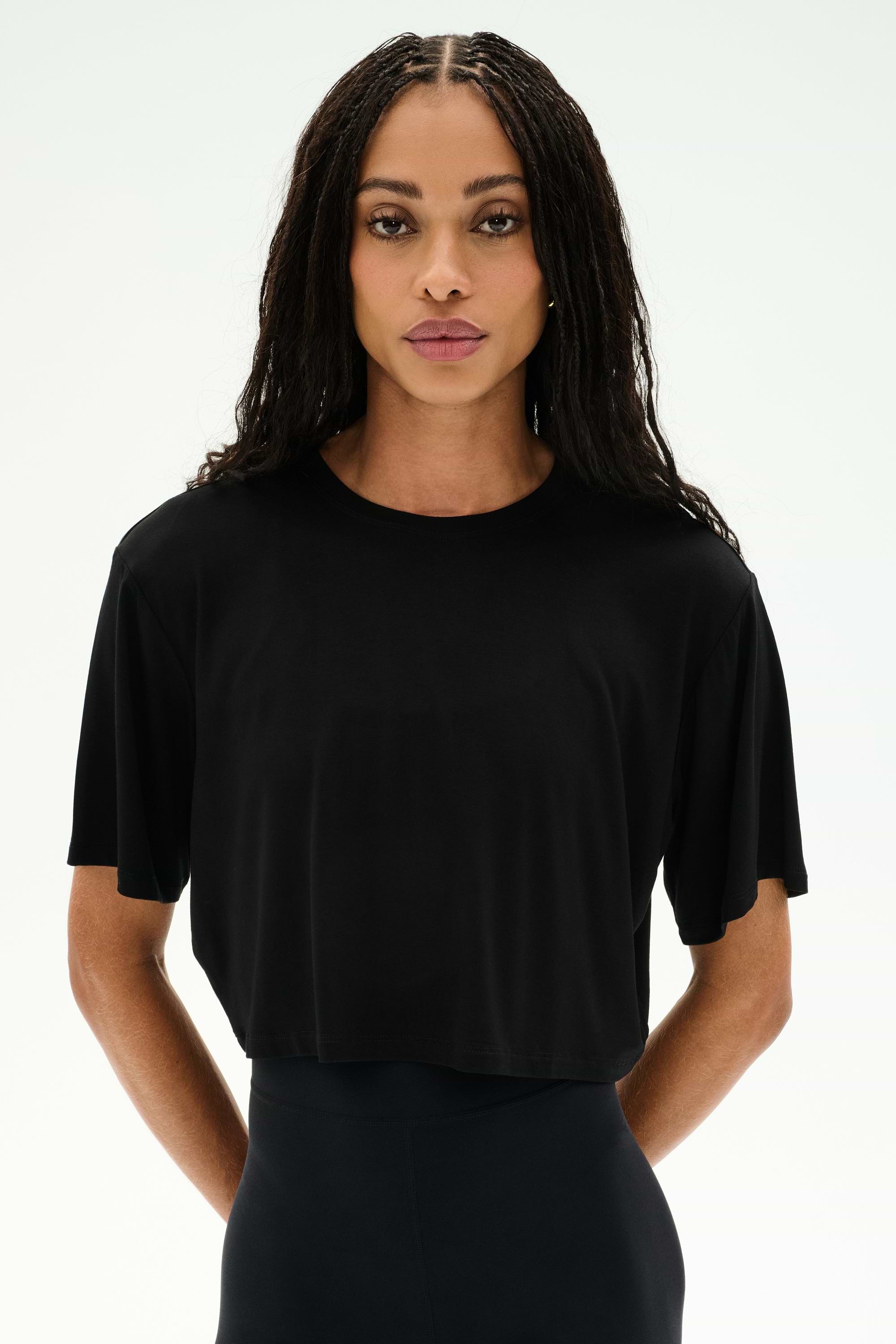 A person with long braided hair wears the SPLITS59 Djuna Oversized Crop Jersey Tee in black and black pants, standing against a plain white background.