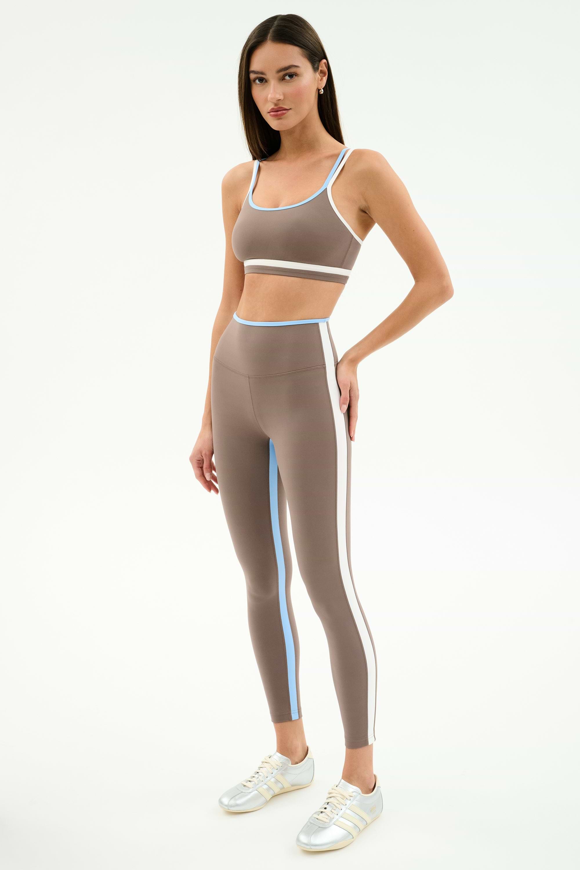 A woman stands in SPLITS59’s Sam High Waist Rigor 7/8 leggings in Lentil/White, featuring compressive fabric and blue and white stripes, paired with a taupe sports bra and white sneakers against a light background.
