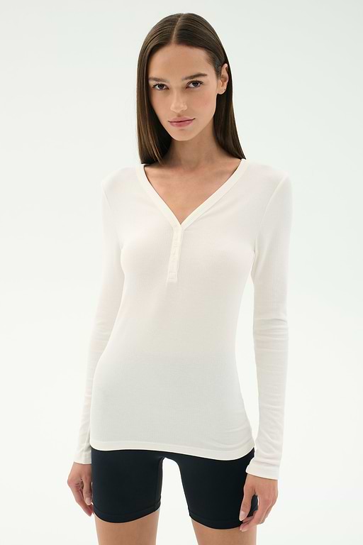 A woman wears the SPLITS59 Louise Rib Henley in white and black spandex shorts, standing against a plain white background.