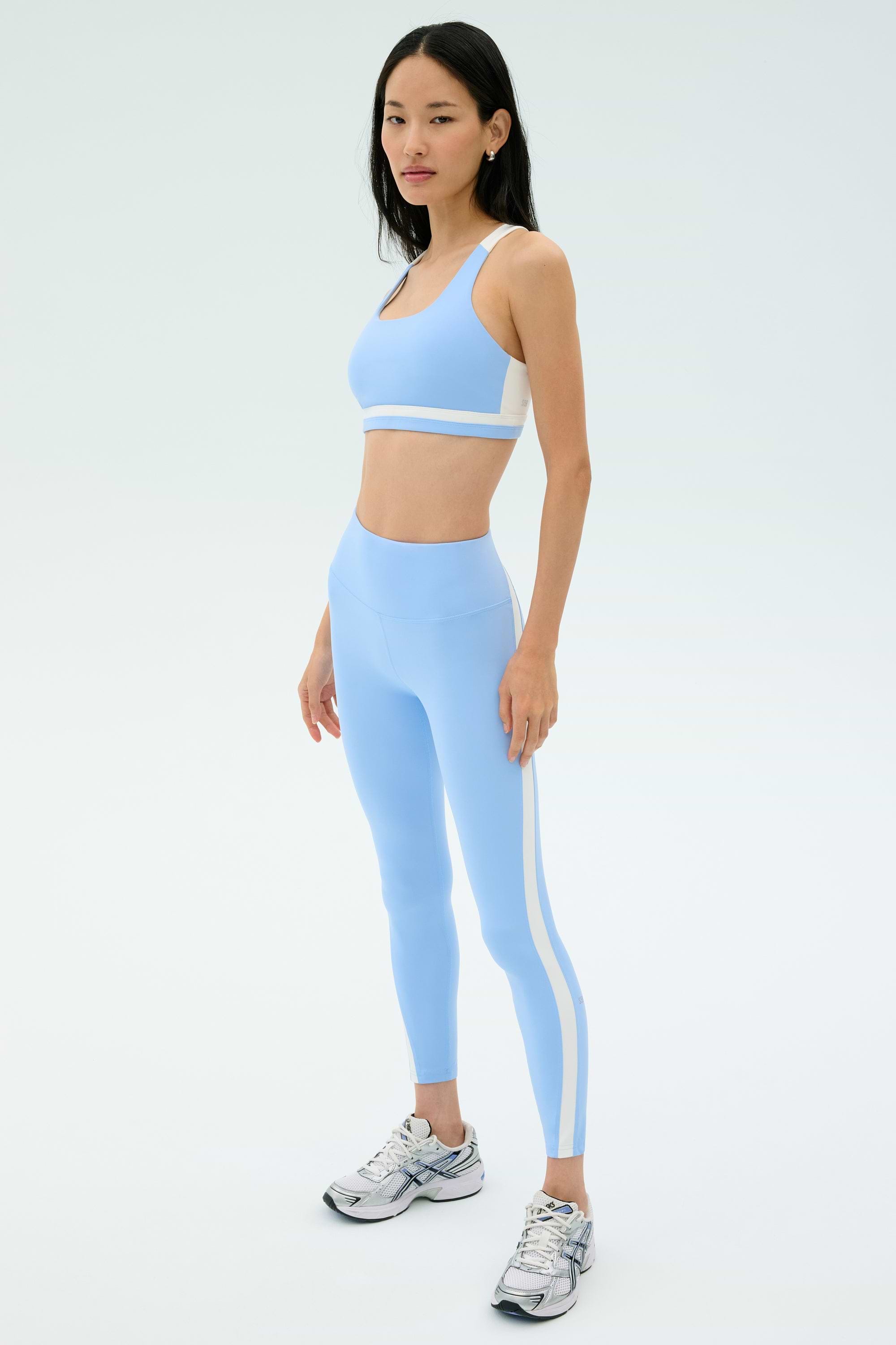 A woman stands on a plain background wearing the SPLITS59 Miles High Waist Rigor 7/8 leggings in Skylight/White and a matching sports bra, paired with silver athletic shoes.