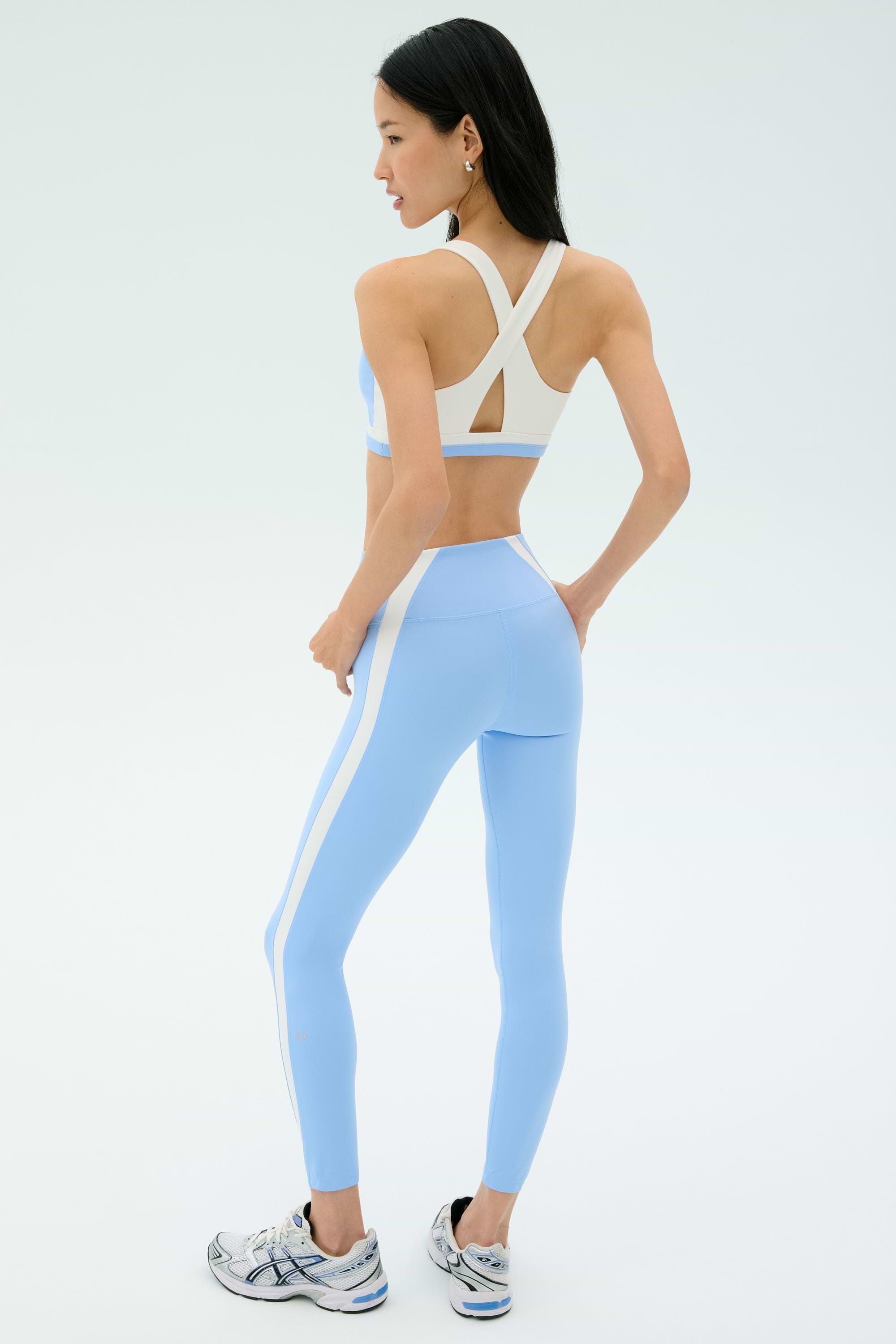 A woman faces away, wearing the SPLITS59 Miles High Waist Rigor 7/8 set in Skylight/White with white sneakers, against a plain light background.