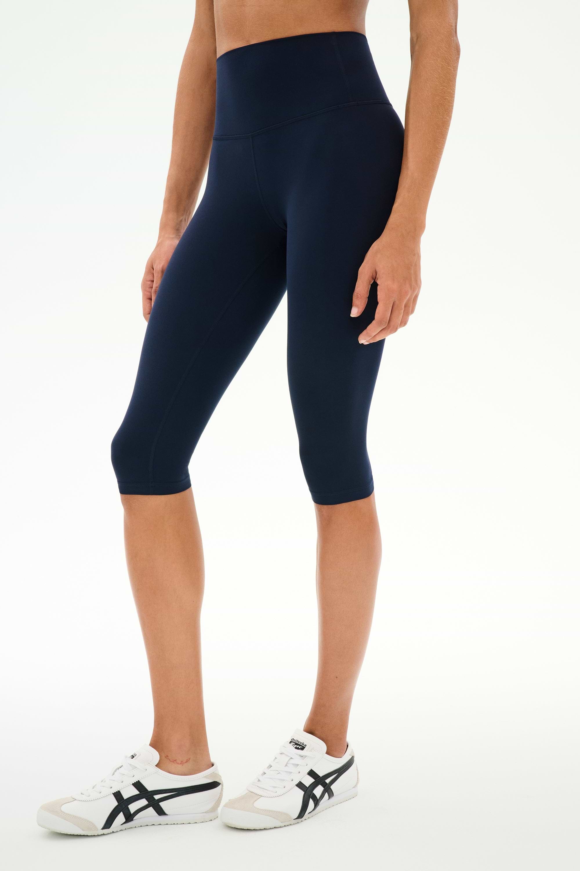 A person wearing the SPLITS59 Airweight High Waist 16" Capri Legging in Indigo and white sneakers, standing against a plain white background.