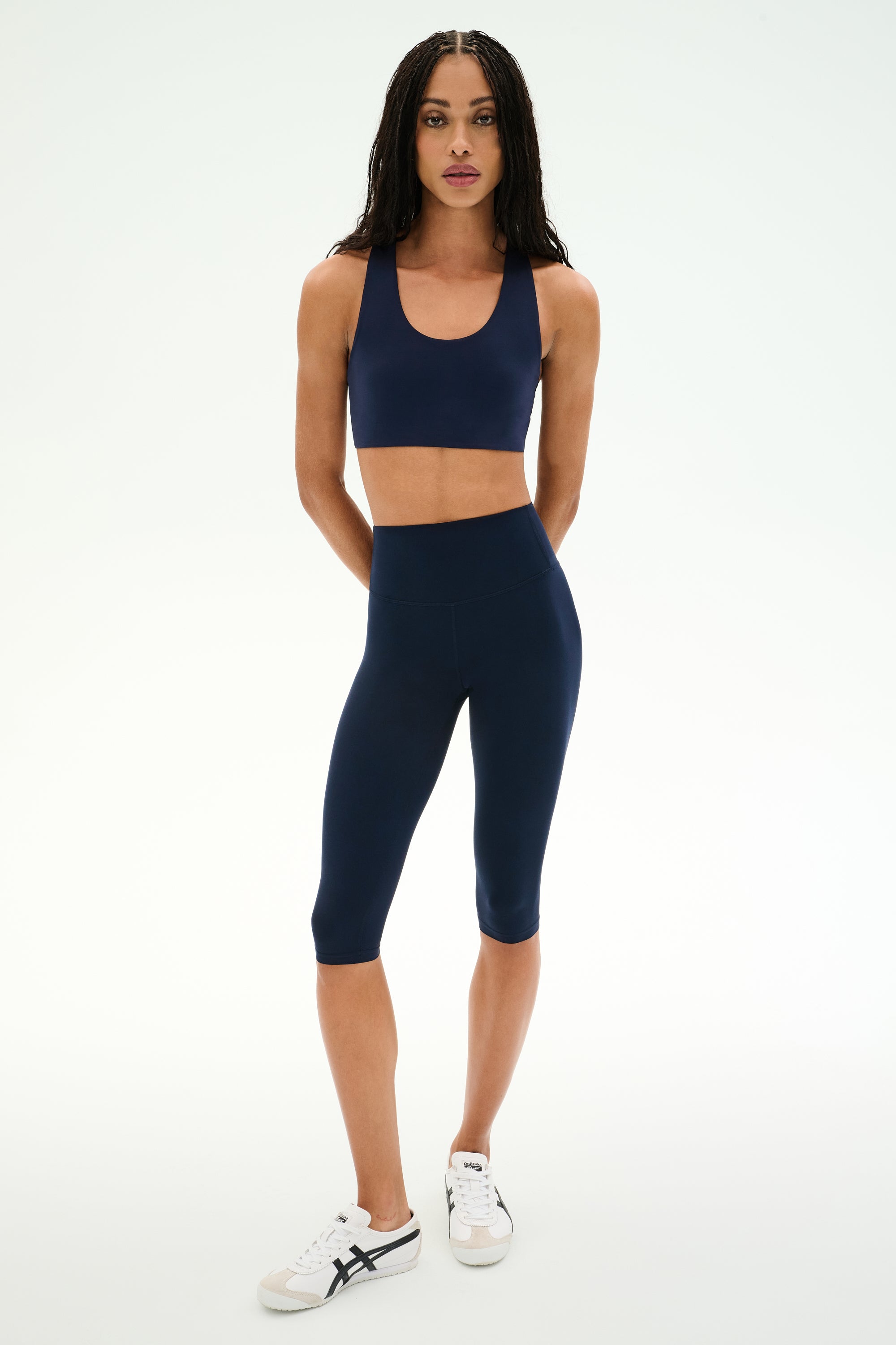 A woman wearing the SPLITS59 Sara Airweight Bra in Indigo with matching knee-length leggings and white sneakers, standing against a plain white background—an ideal outfit for hot yoga or Pilates sessions.