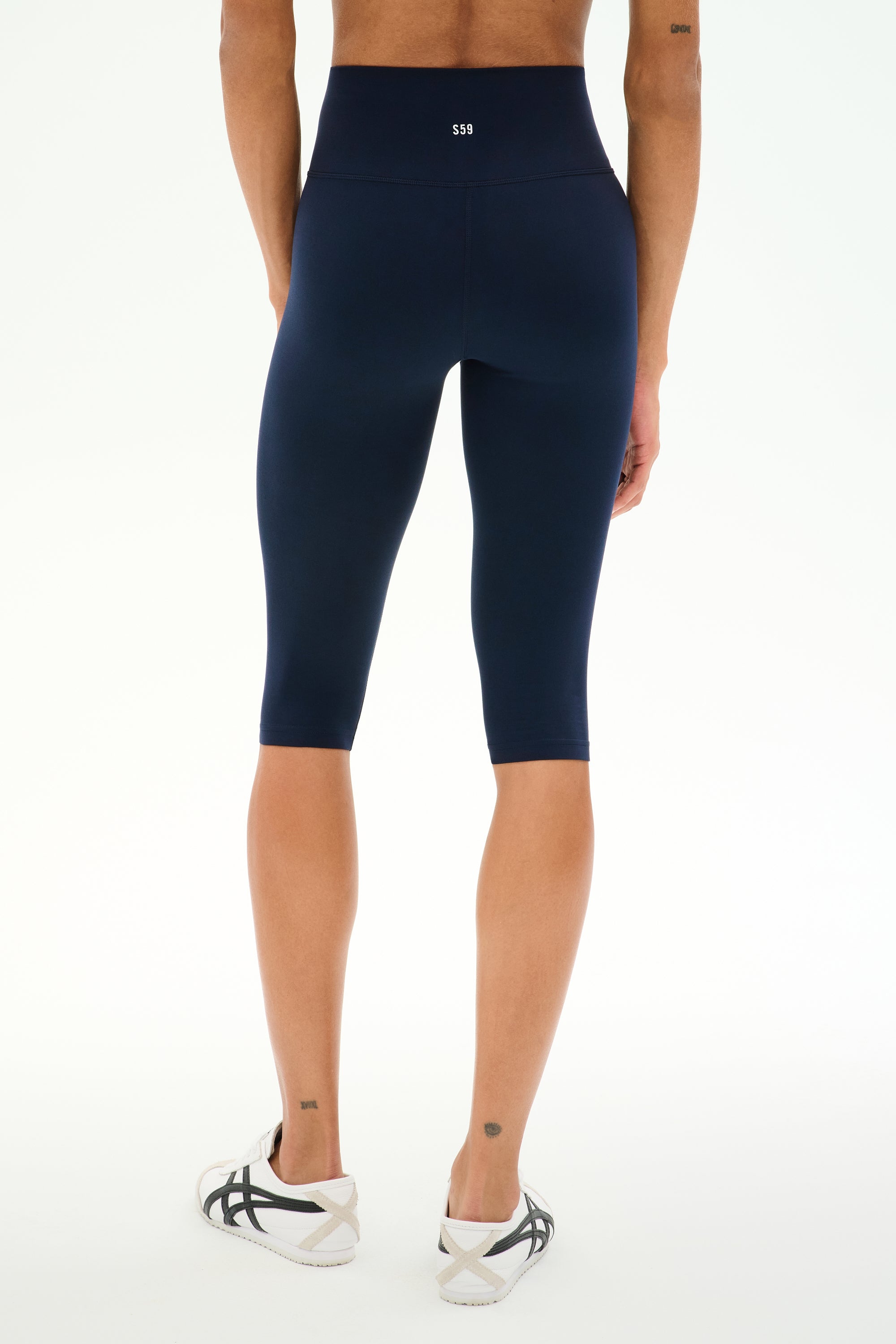 A person is wearing SPLITS59 Airweight High Waist 16" Capri Legging in Indigo and white sneakers, standing on a white background, viewed from the back.