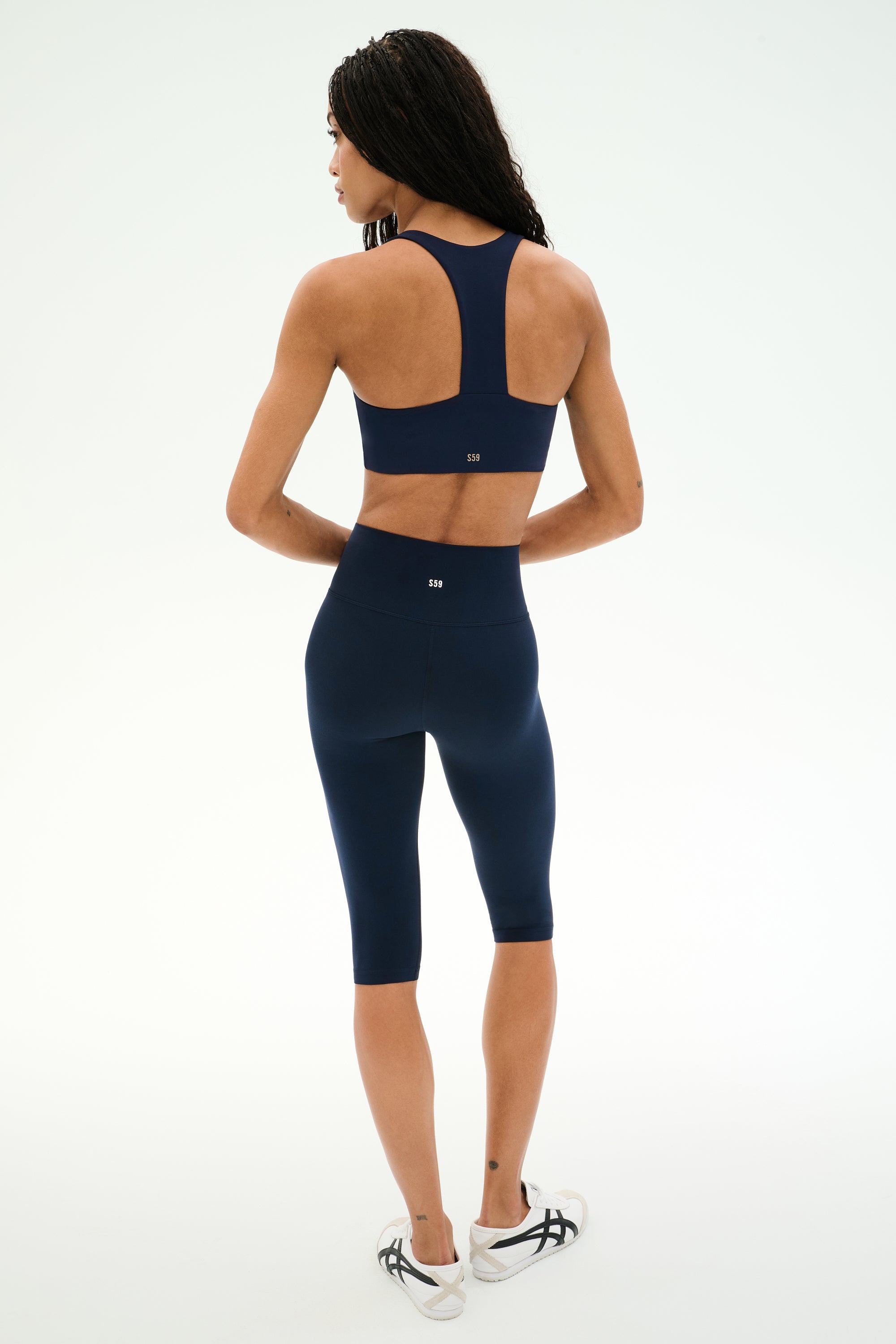 A woman stands facing away, wearing the SPLITS59 Sara Airweight Bra in Indigo and matching high-waisted biker shorts—ideal for Pilates or hot yoga—paired with white athletic shoes, set against a plain light background.