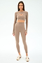A woman faces forward wearing the SPLITS59 Amelia Airweight Lite Shrug in Latte—a taupe, long-sleeve crop top ideal for yoga and dance—styled with matching high-waisted leggings and beige sneakers.
