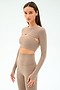 A woman wears the SPLITS59 Amelia Airweight Lite Shrug in Latte, a beige, long-sleeve crop top with a cutout detail, paired with matching high-waisted leggings—ideal for yoga or dance warmups against a plain background.