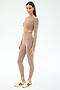 A woman stands in a taupe athletic set with long sleeves and high-waisted leggings, cream and maroon sneakers, and the SPLITS59 Amelia Airweight Lite Shrug - Latte—perfect for yoga or dance warmups—on a white background.