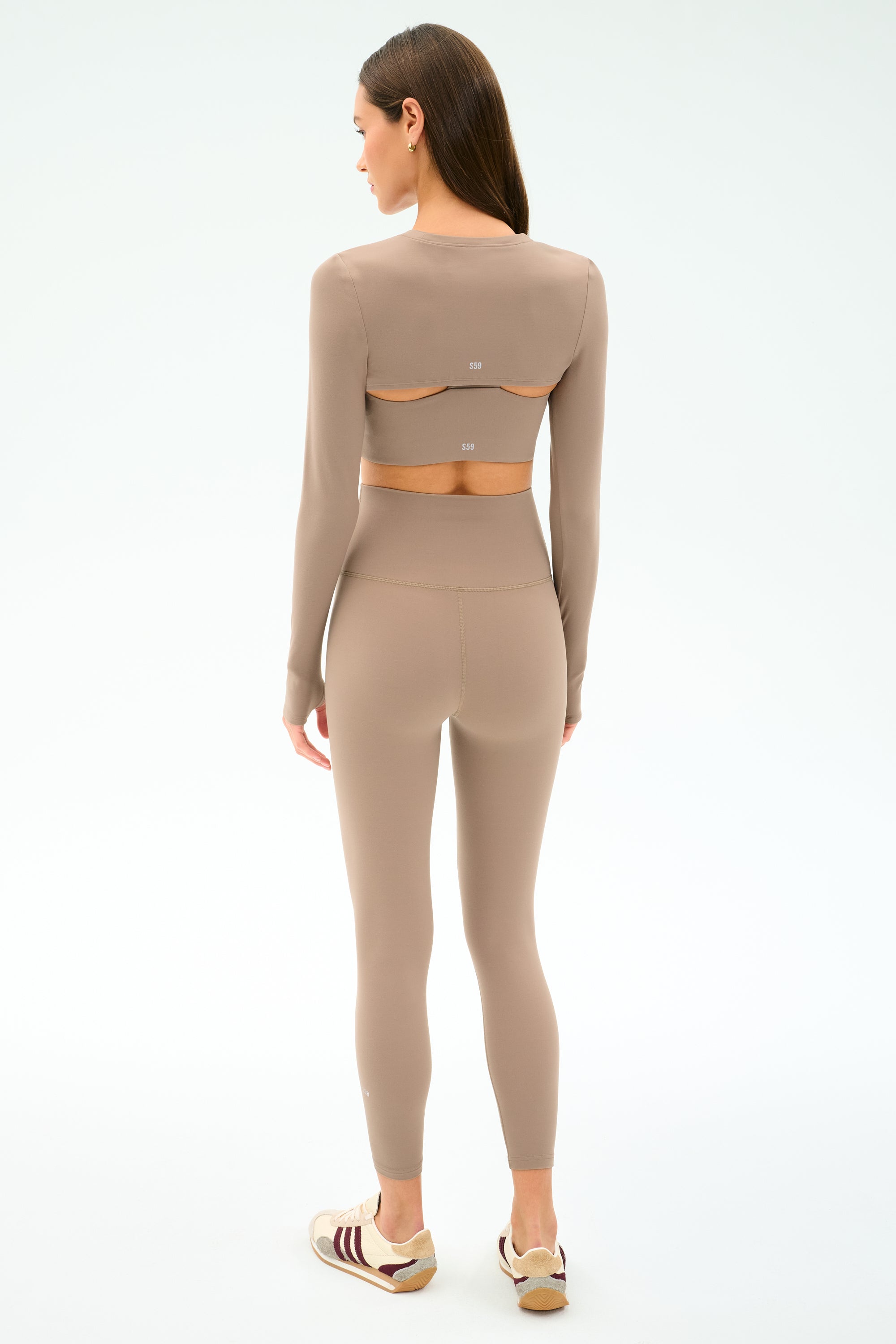 A woman faces away, wearing the SPLITS59 Amelia Airweight Lite Shrug in Latte—ideal for dance warmups or yoga—with white and maroon sneakers against a plain light background.