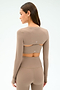 A woman seen from the back wears the SPLITS59 Amelia Airweight Lite Shrug in Latte, a taupe long-sleeve fitted top with cutout details and small printed numbers—ideal for yoga or dance warmups.