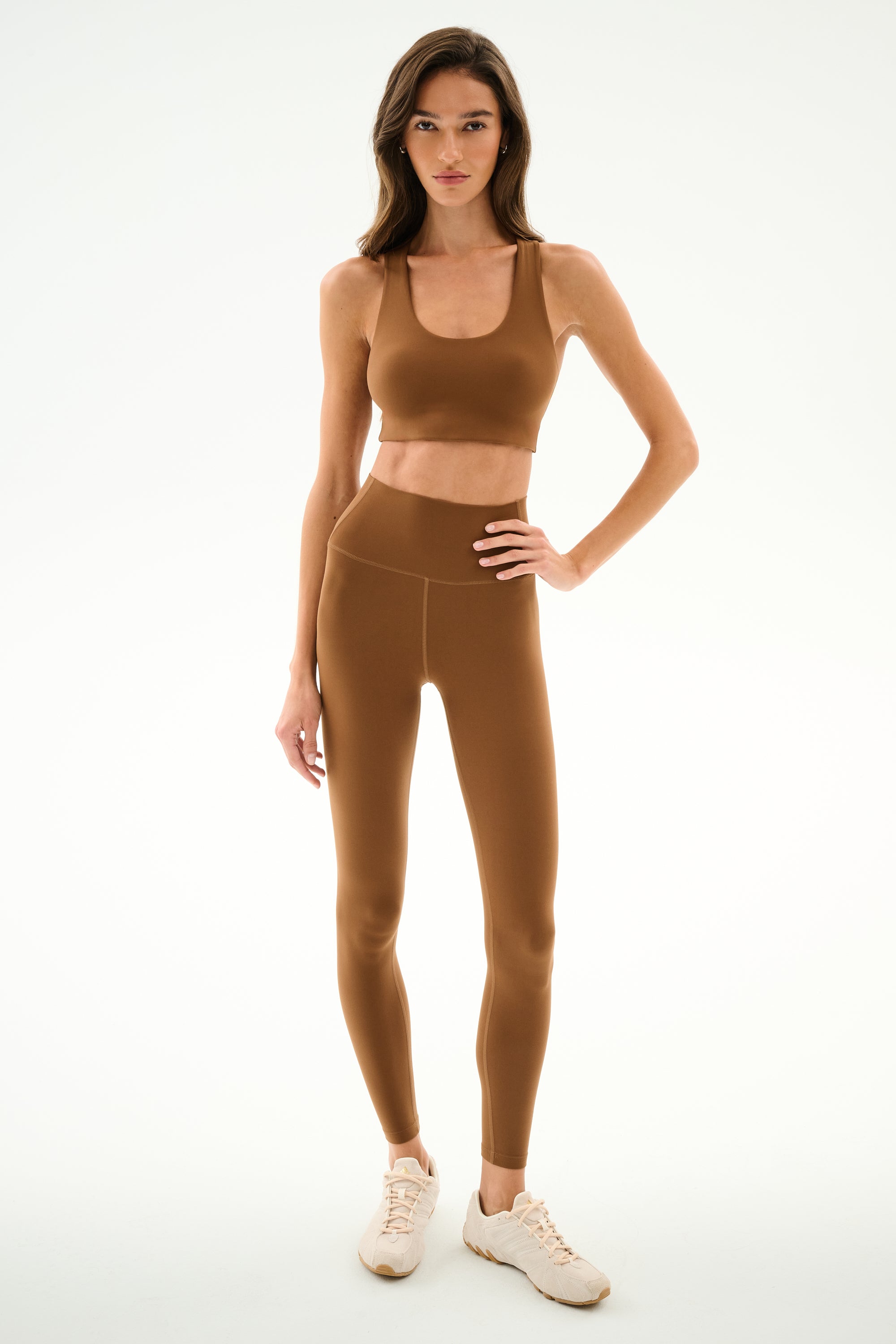 Woman wearing SPLITS59 Airweight High Waist - Toffee leggings and sports bra with light sneakers, standing against a plain white background.