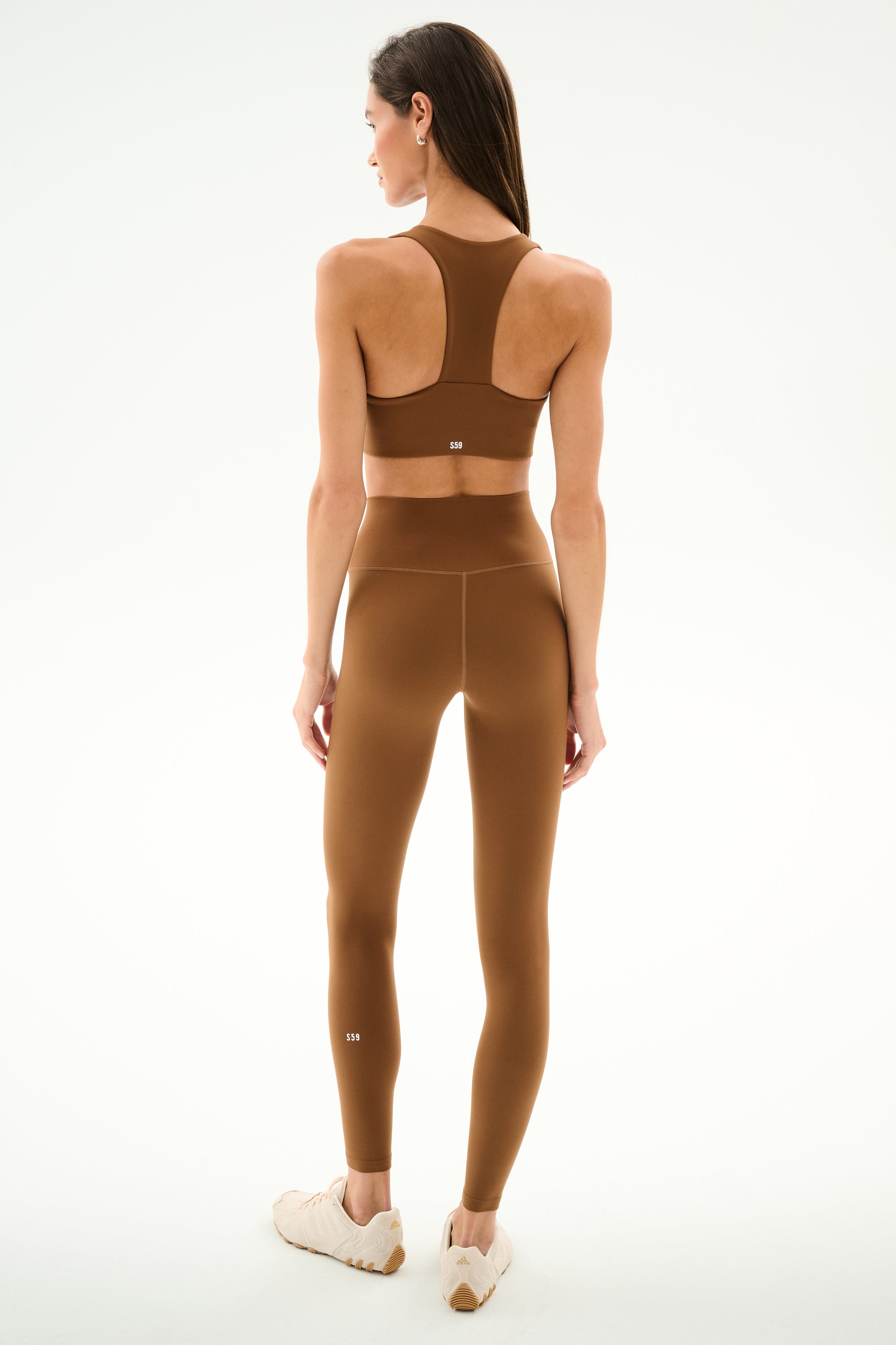Woman stands facing away in SPLITS59 Airweight High Waist - Toffee leggings and sports bra with beige sneakers, ideal for hot yoga, on a plain white background.