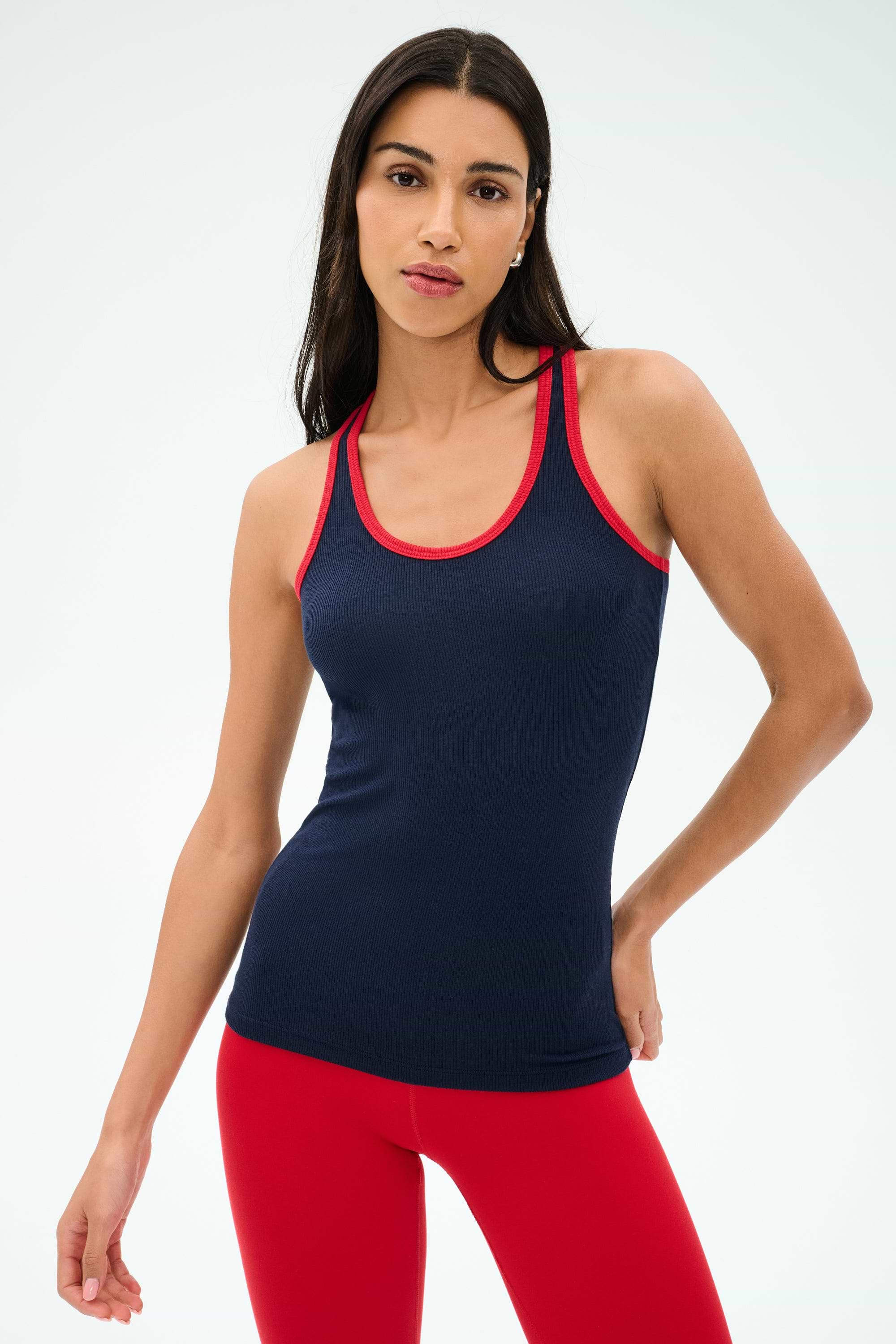 A woman in the SPLITS59 Ashby Rib Tank - Indigo/Pirate Red stands against a plain light background, wearing red leggings and ready for yoga or gym workouts.