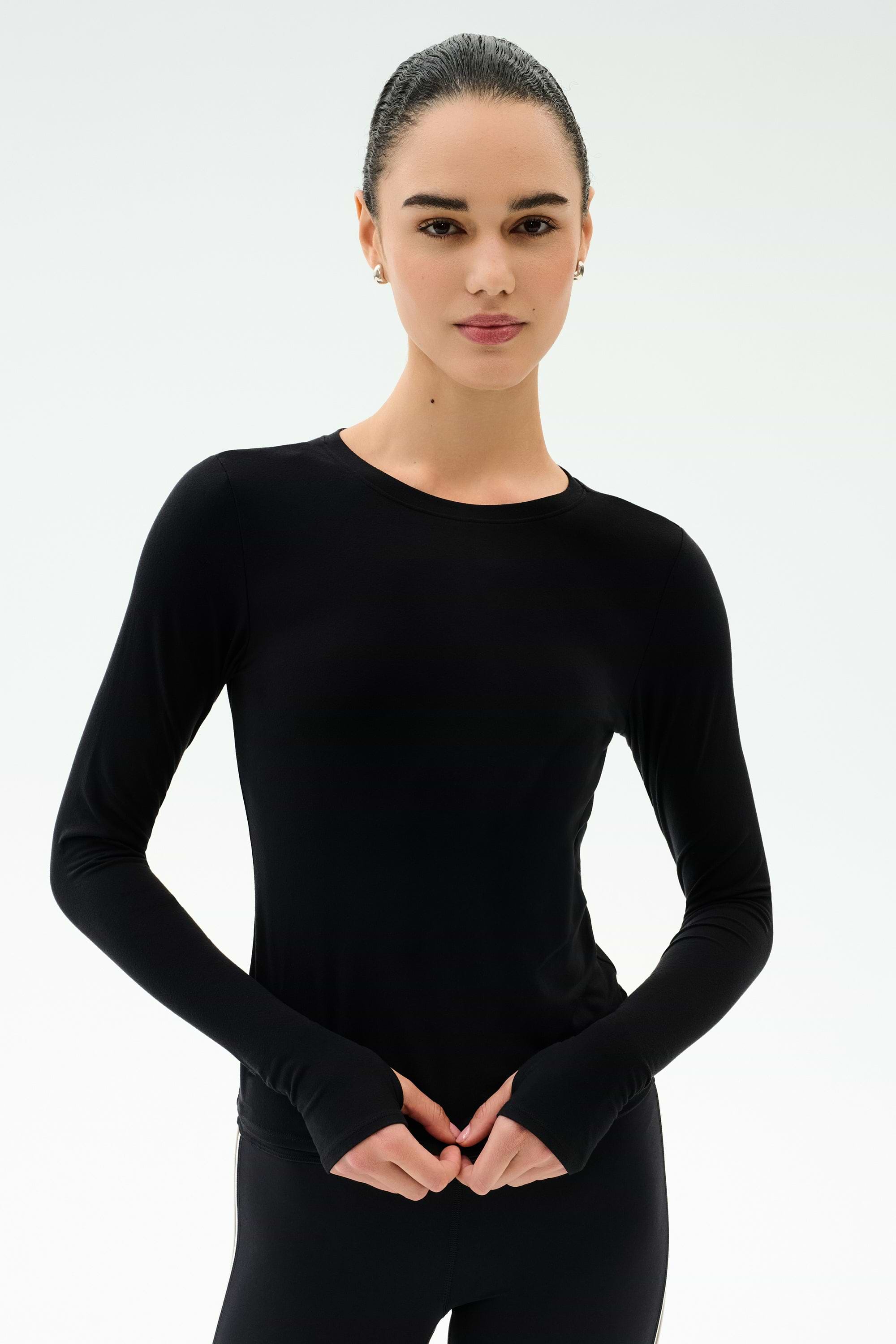 A woman with dark hair tied back wears the SPLITS59 Airweight Lite Long Sleeve Waist Length top in black, paired with black pants and posed against a plain white background—perfect for gym workouts.