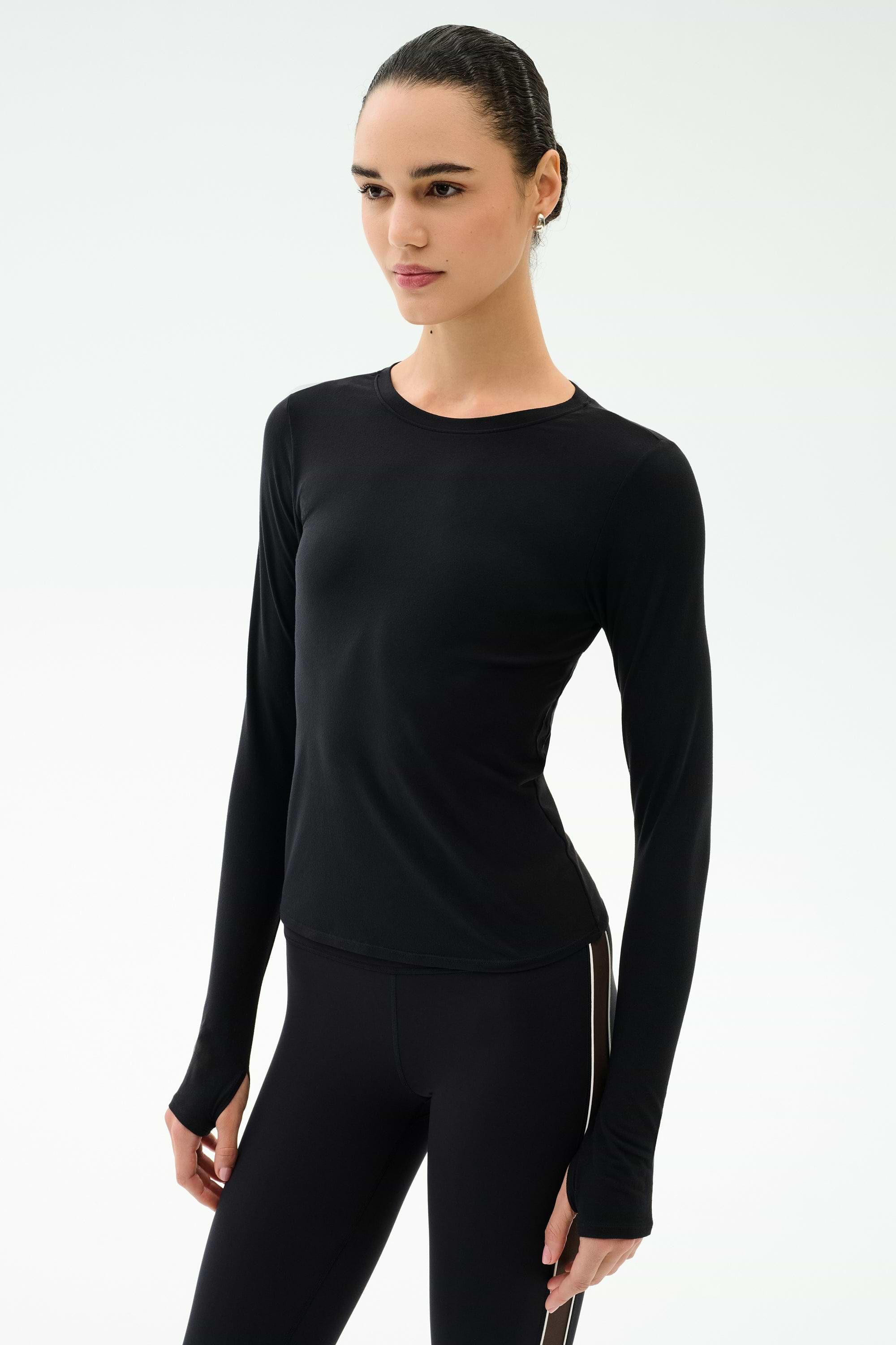 A woman wears the SPLITS59 Airweight Lite Long Sleeve Waist Length top in black, paired with black leggings, standing against a plain background—the quick-dry comfort is perfect for gym workouts.