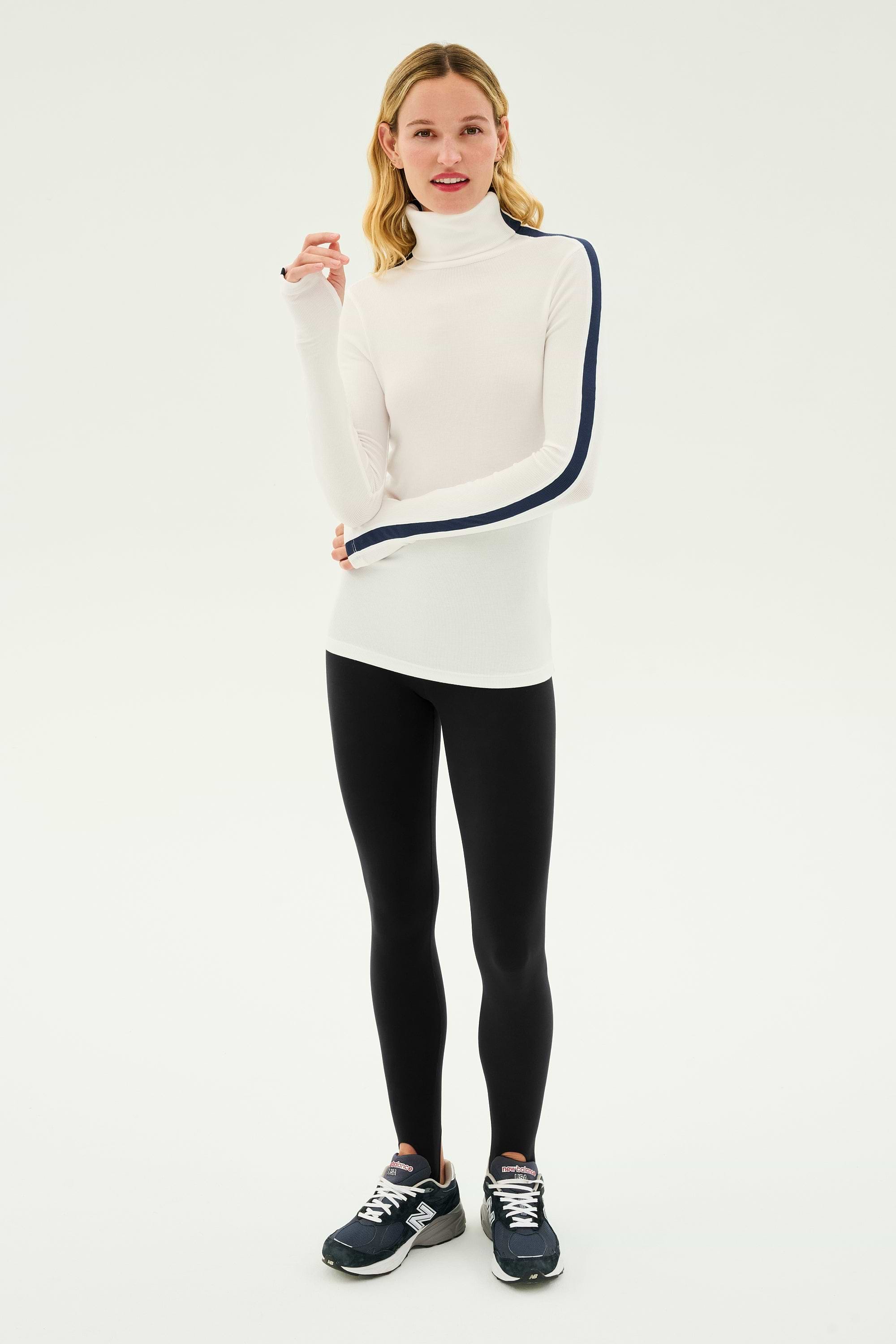 A person wearing a SPLITS59 Jackson Rib Full Length Turtleneck in White/Indigo, black leggings, and black athletic shoes stands against a plain background.