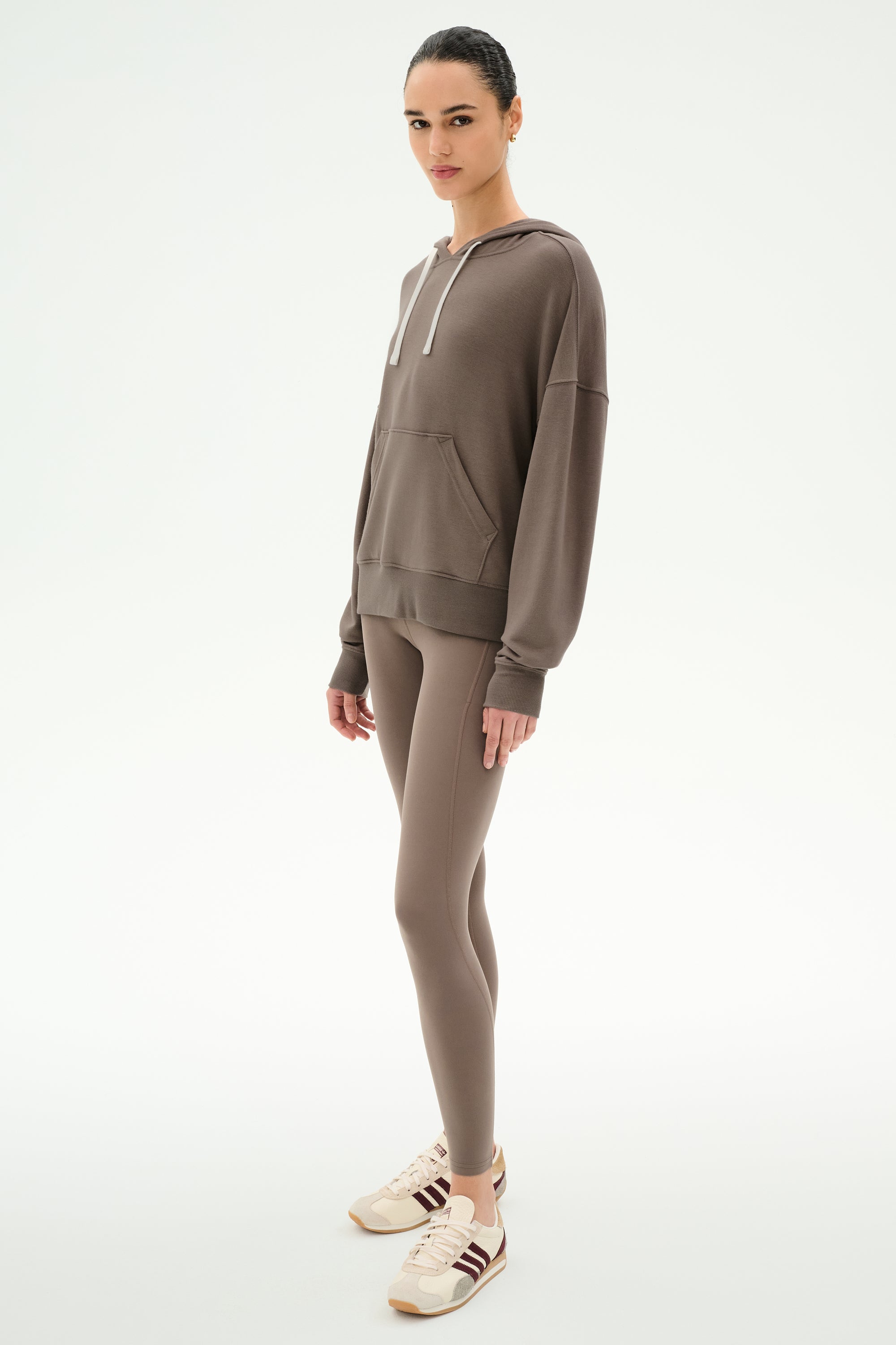 A person stands against a white background, wearing the SPLITS59 Joey Fleece Hoodie in Lentil, paired with matching brown leggings and beige sneakers featuring brown stripes.