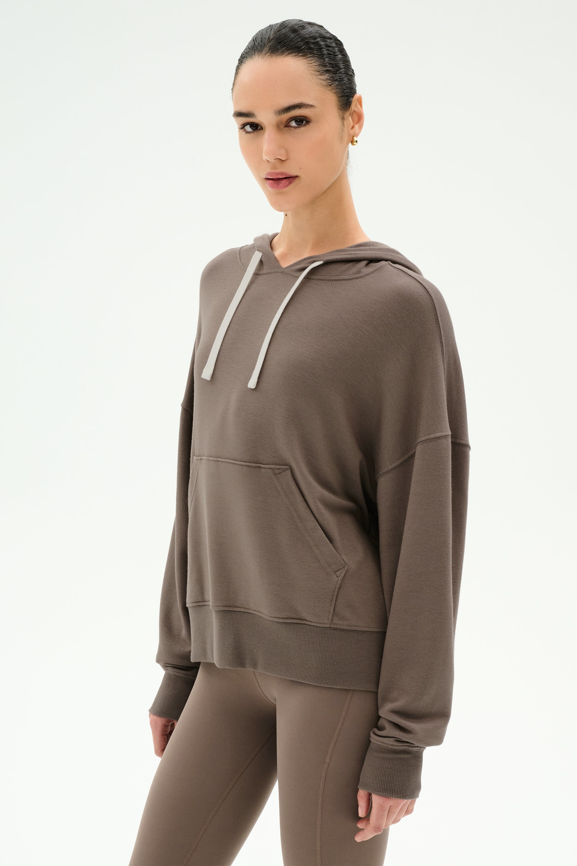 A woman stands against a plain white background wearing the SPLITS59 Joey Fleece Hoodie in Lentil, crafted from an ultra-soft modal spandex blend with a front pocket, paired with matching brown leggings.