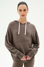 A woman wears the SPLITS59 Joey Fleece Hoodie in Lentil—a brown hoodie crafted from a soft modal spandex blend with white drawstrings—standing against a plain light background and looking directly at the camera with her hands clasped in front.