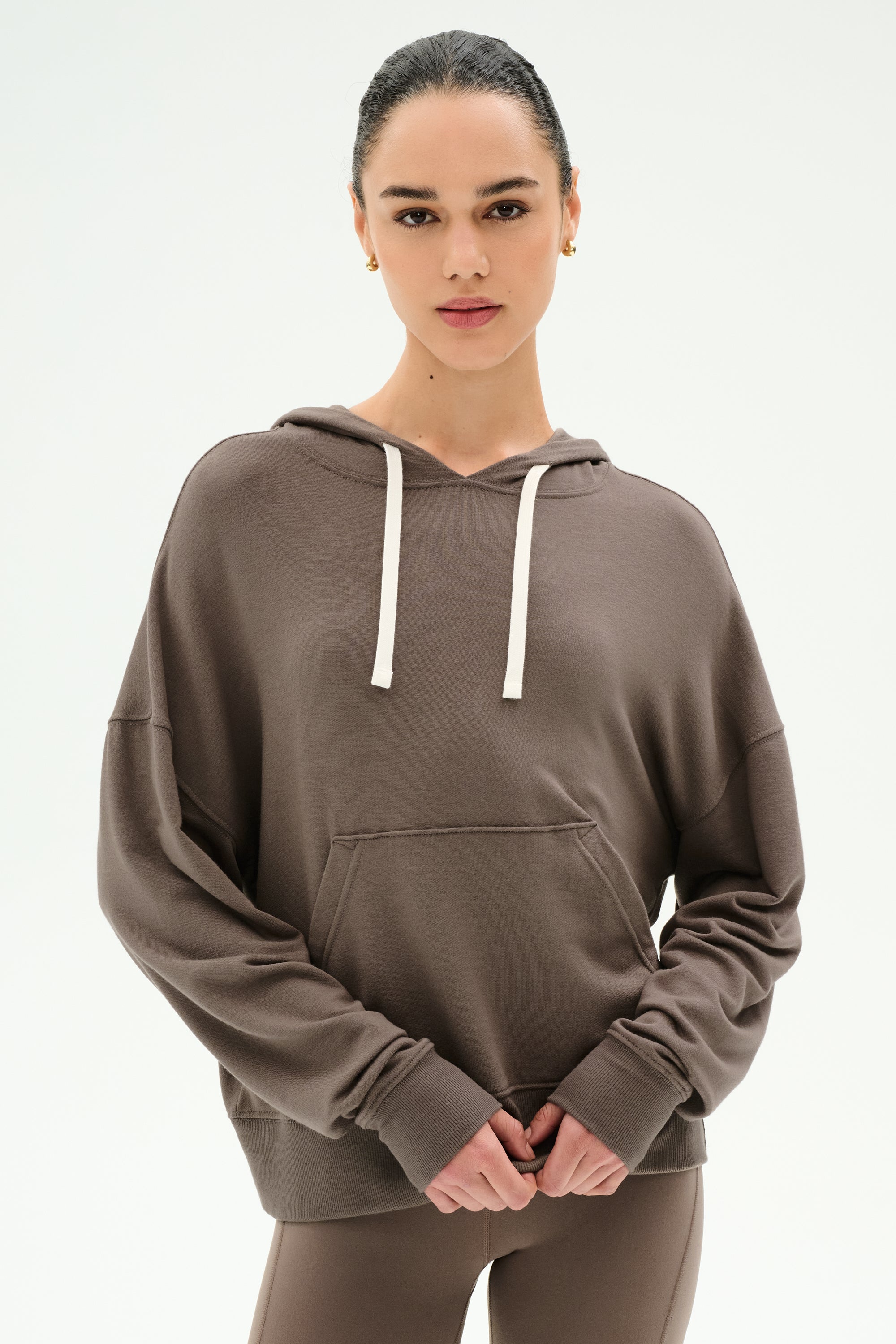 A woman wears the SPLITS59 Joey Fleece Hoodie in Lentil—a brown hoodie crafted from a soft modal spandex blend with white drawstrings—standing against a plain light background and looking directly at the camera with her hands clasped in front.