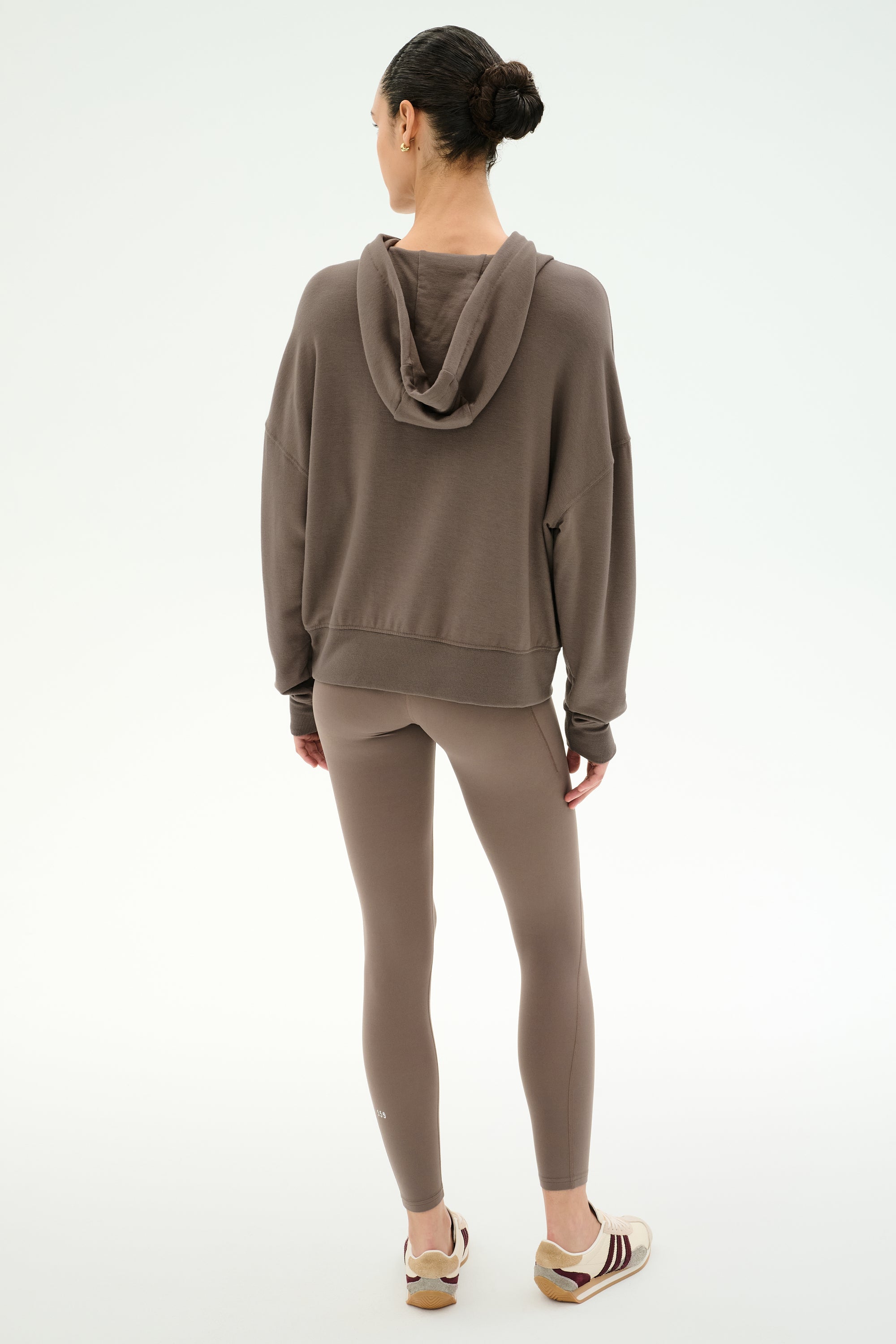 A person stands facing away, dressed in the SPLITS59 Joey Fleece Hoodie in Lentil—a brown hooded sweatshirt crafted from a soft modal spandex blend—paired with matching leggings and beige sneakers with stripes, set against a plain light background.