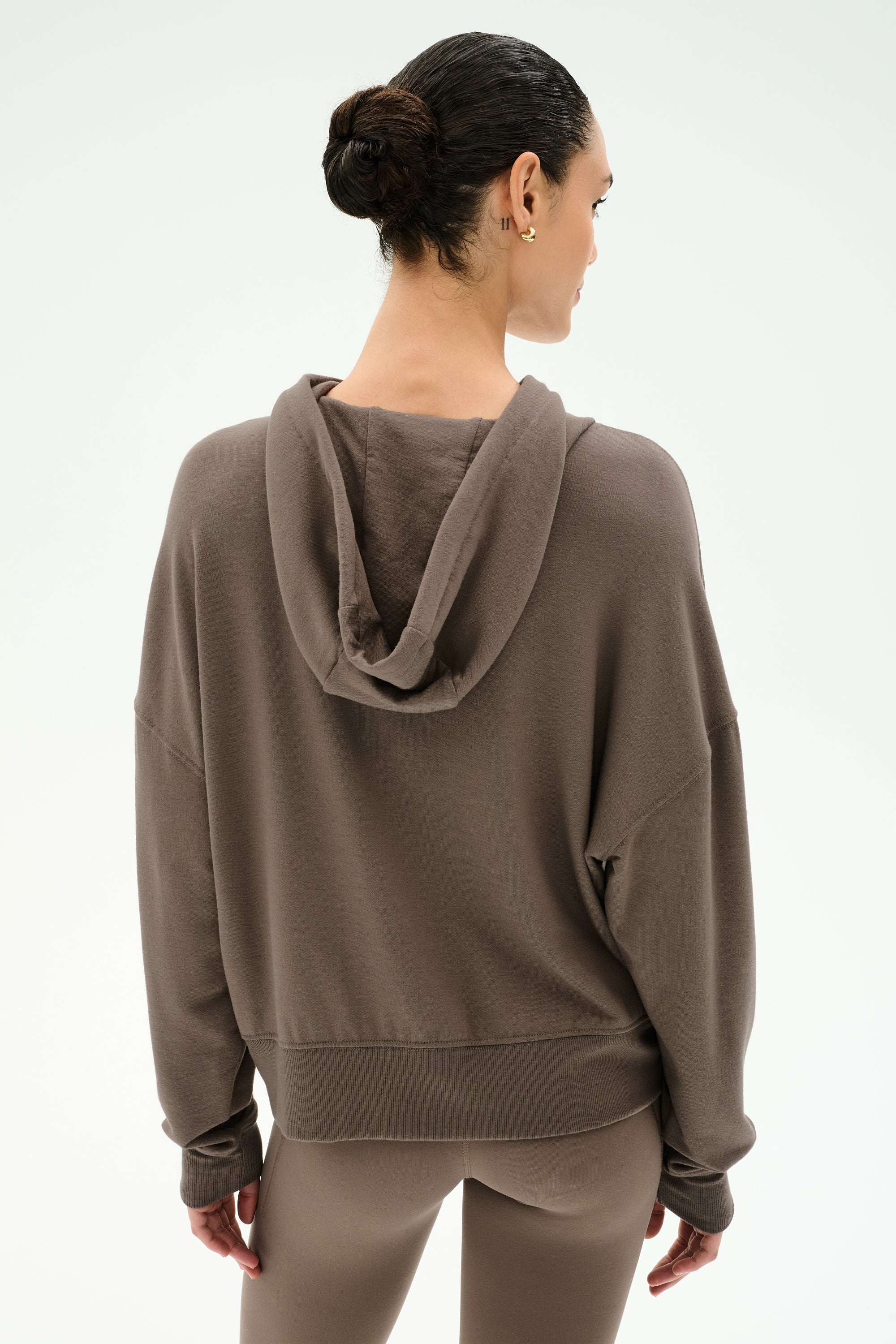 A woman with dark hair in a bun wears the SPLITS59 Joey Fleece Hoodie in Lentil and matching leggings, crafted from a soft modal spandex blend, facing away from the camera against a plain light background.