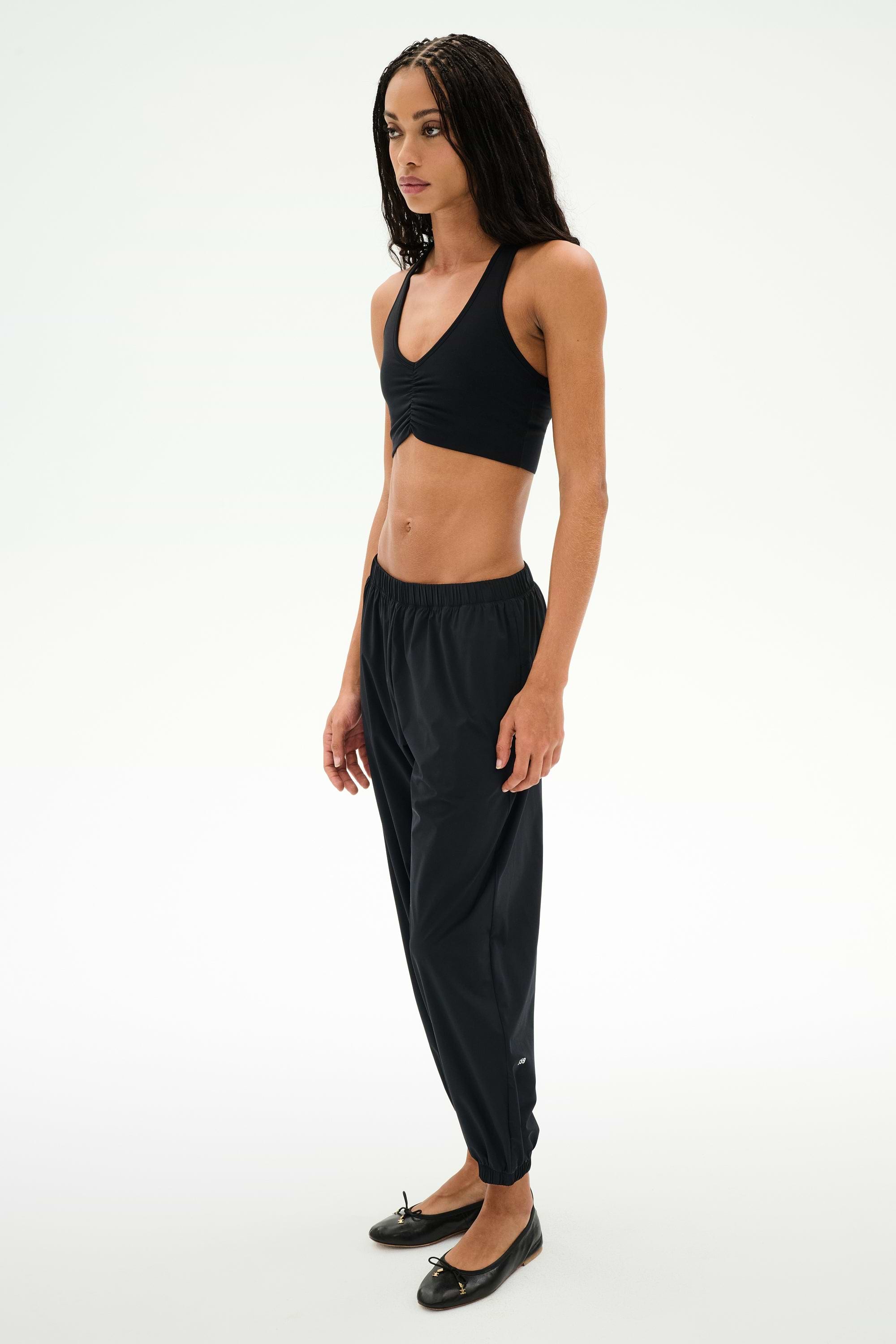 A person stands against a plain background wearing a black crop top, black SPLITS59 Megan Woven Nylon Jogger pants with a cinched waistband, and black ballet flats.
