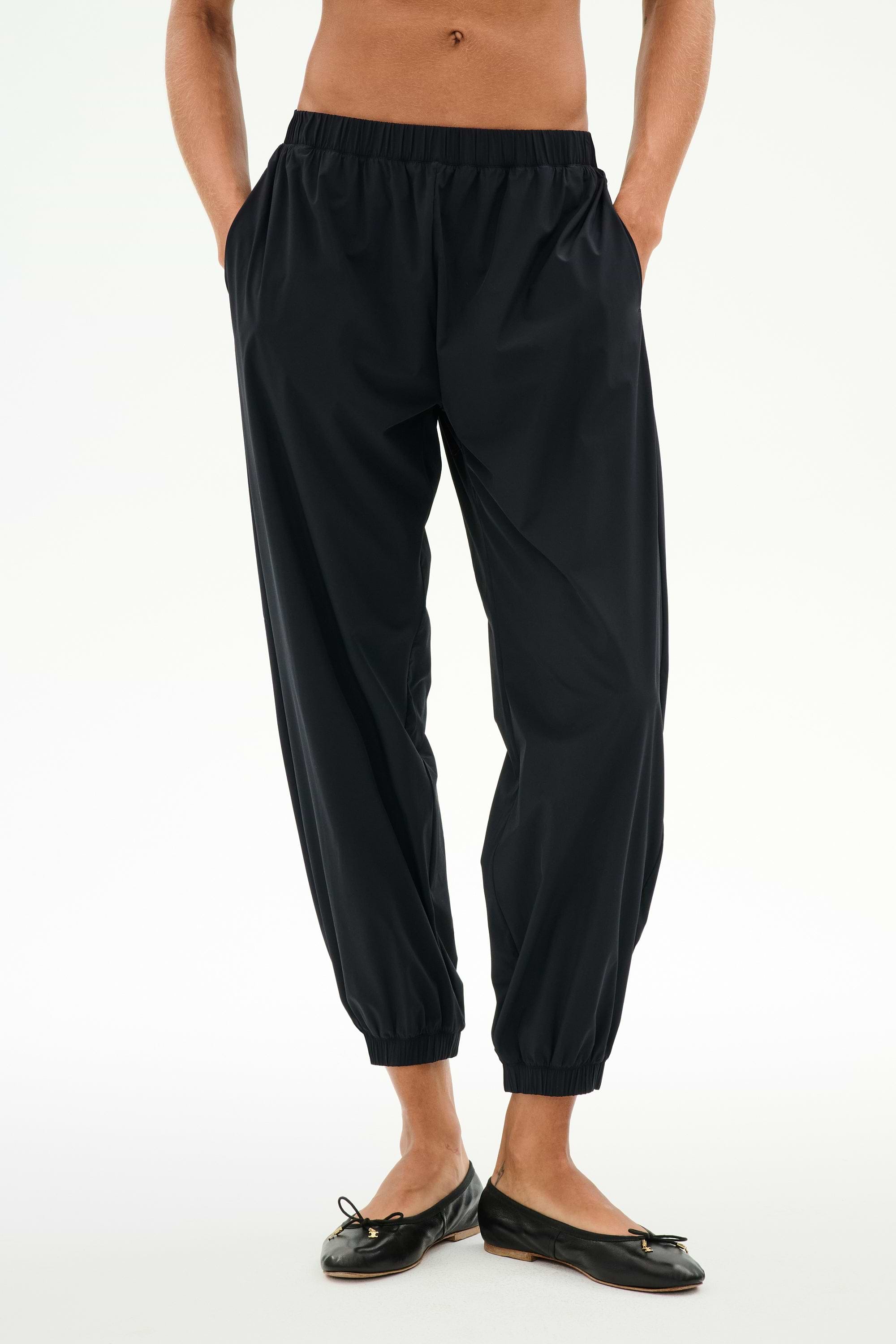 Model wears the SPLITS59 Megan Woven Nylon Jogger in black, featuring a cinched waistband and hands in pockets, paired with black ballet flats. Shown from chest down against a plain background.