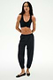 A woman stands against a white background wearing SPLITS59's Megan Woven Nylon Jogger in black, paired with a black sports bra and black flats, her hands behind her back.