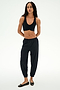A woman stands against a white background wearing SPLITS59's Megan Woven Nylon Jogger in black, paired with a black sports bra and black flats, her hands behind her back.