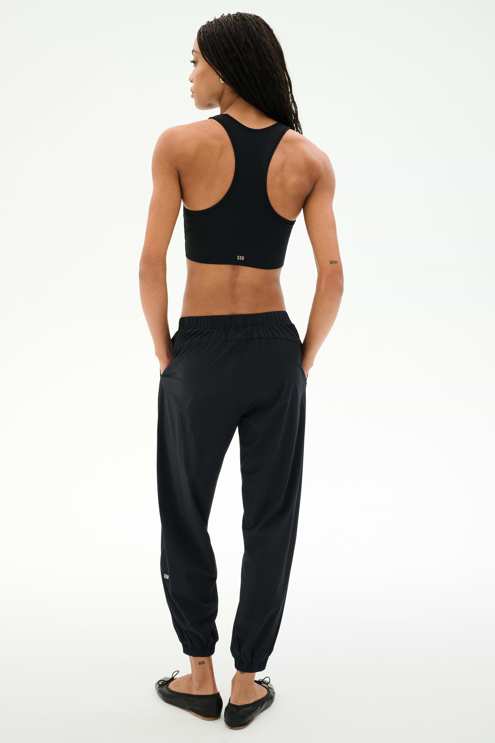 Person standing with their back to the camera, wearing SPLITS59’s Megan Woven Nylon Jogger - Black, a racerback sports top, and black shoes against a plain light background.