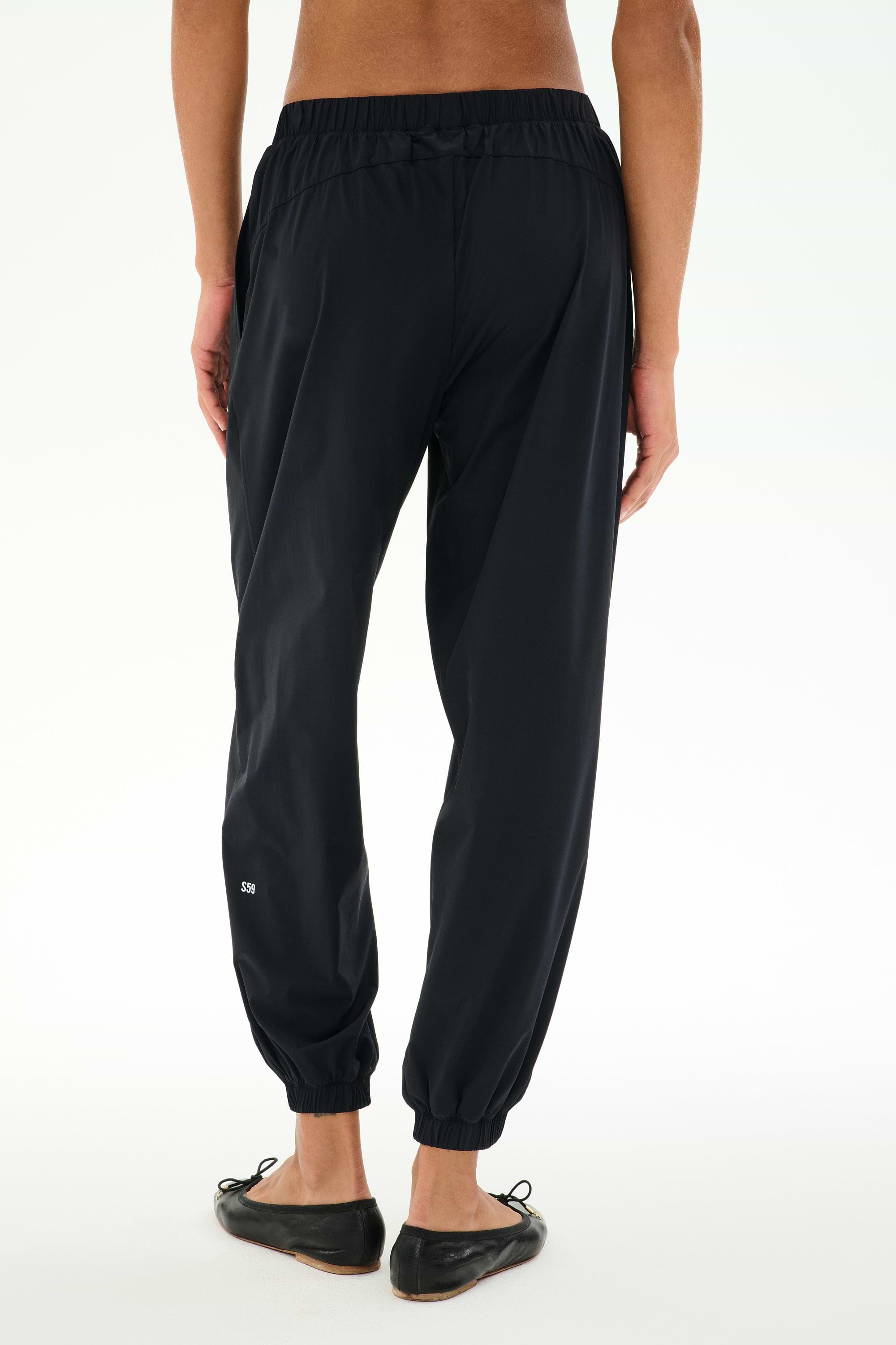 A person is seen from behind, standing against a white background, wearing SPLITS59 Megan Woven Nylon Jogger in black paired with black flat shoes.