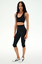 A woman wearing the SPLITS59 Sara Airweight Bra in black, paired with black high-waisted capri leggings and white athletic shoes, stands against a plain white background—an ideal outfit for Pilates or hot yoga sessions.