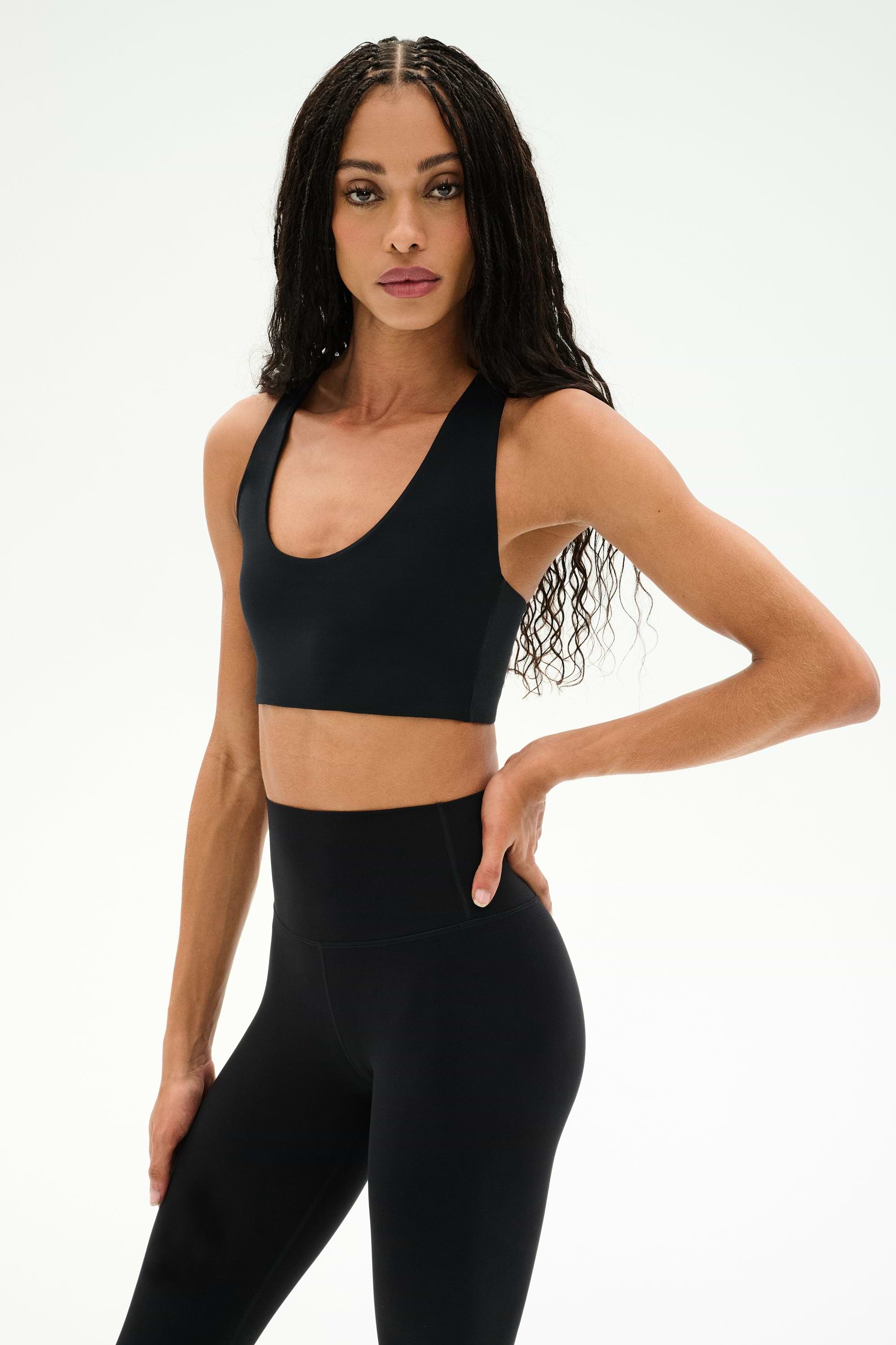 A woman with long braided hair, dressed in the SPLITS59 Sara Airweight Bra in black and high-waisted leggings, stands against a plain light background facing forward with one hand on her hip—perfectly outfitted for pilates or hot yoga.