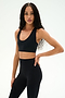 A woman with long braided hair, dressed in the SPLITS59 Sara Airweight Bra in black and high-waisted leggings, stands against a plain light background facing forward with one hand on her hip—perfectly outfitted for pilates or hot yoga.