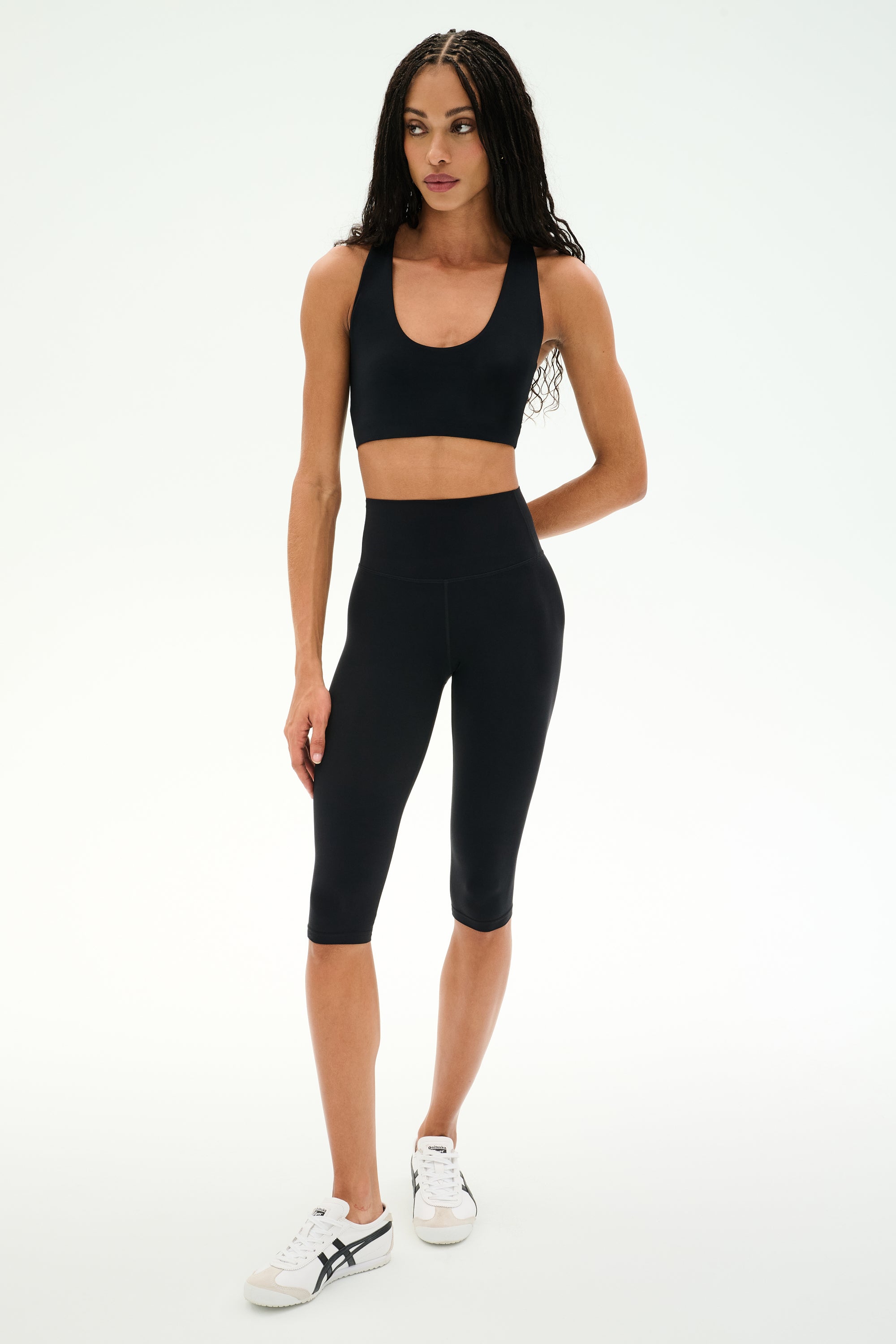 A woman stands against a white background, wearing a black sports bra, SPLITS59 Airweight High Waist 16" Capri Legging in black, and white sneakers.