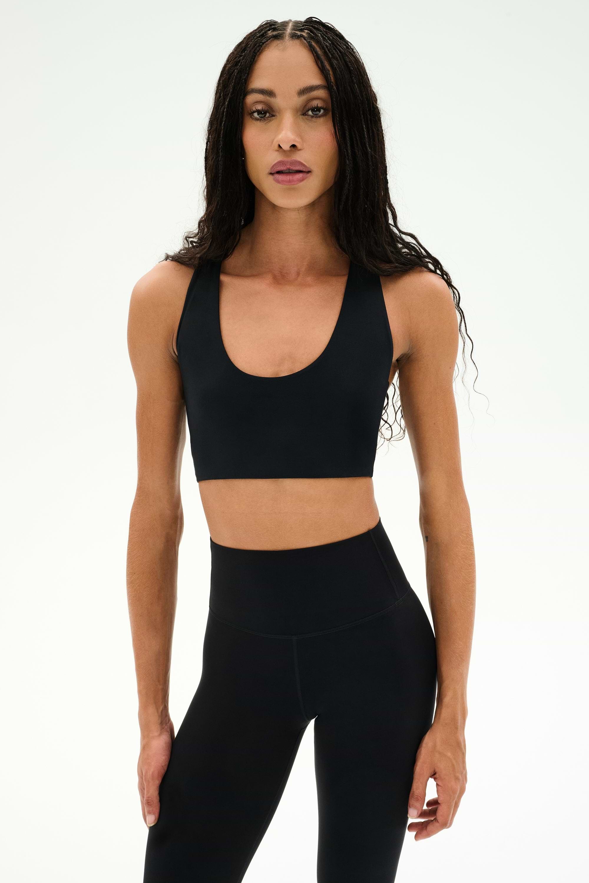 Wearing the SPLITS59 Sara Airweight Bra in black and matching leggings, a person with long braided hair stands against a plain white background—an ideal outfit for hot yoga or Pilates.