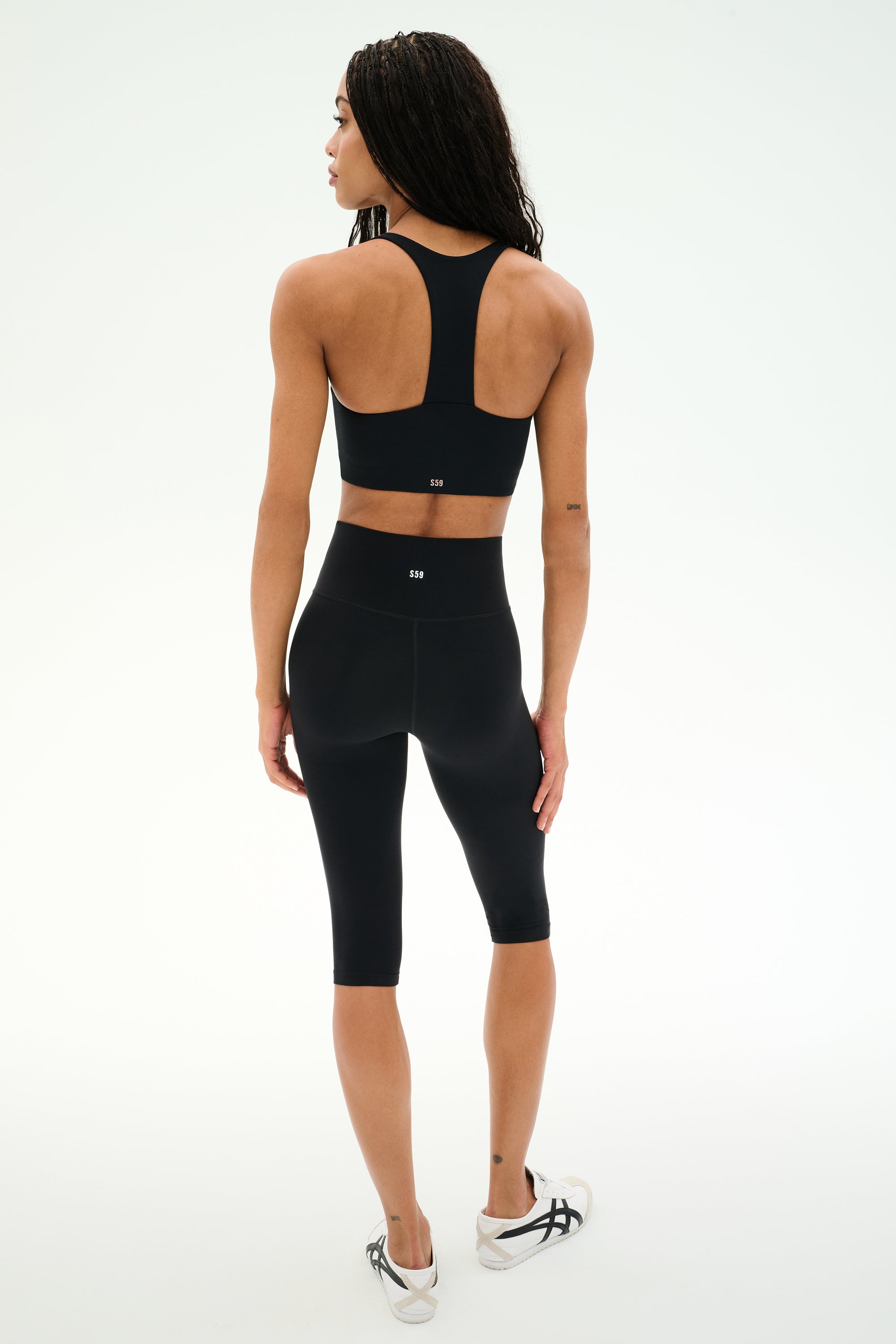 A person stands on a white background, facing away from the camera, wearing the SPLITS59 Sara Airweight Bra in black paired with black high-waisted knee-length leggings ideal for hot yoga or Pilates, and white sneakers.