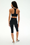 A person stands on a white background, facing away from the camera, wearing the SPLITS59 Sara Airweight Bra in black paired with black high-waisted knee-length leggings ideal for hot yoga or Pilates, and white sneakers.