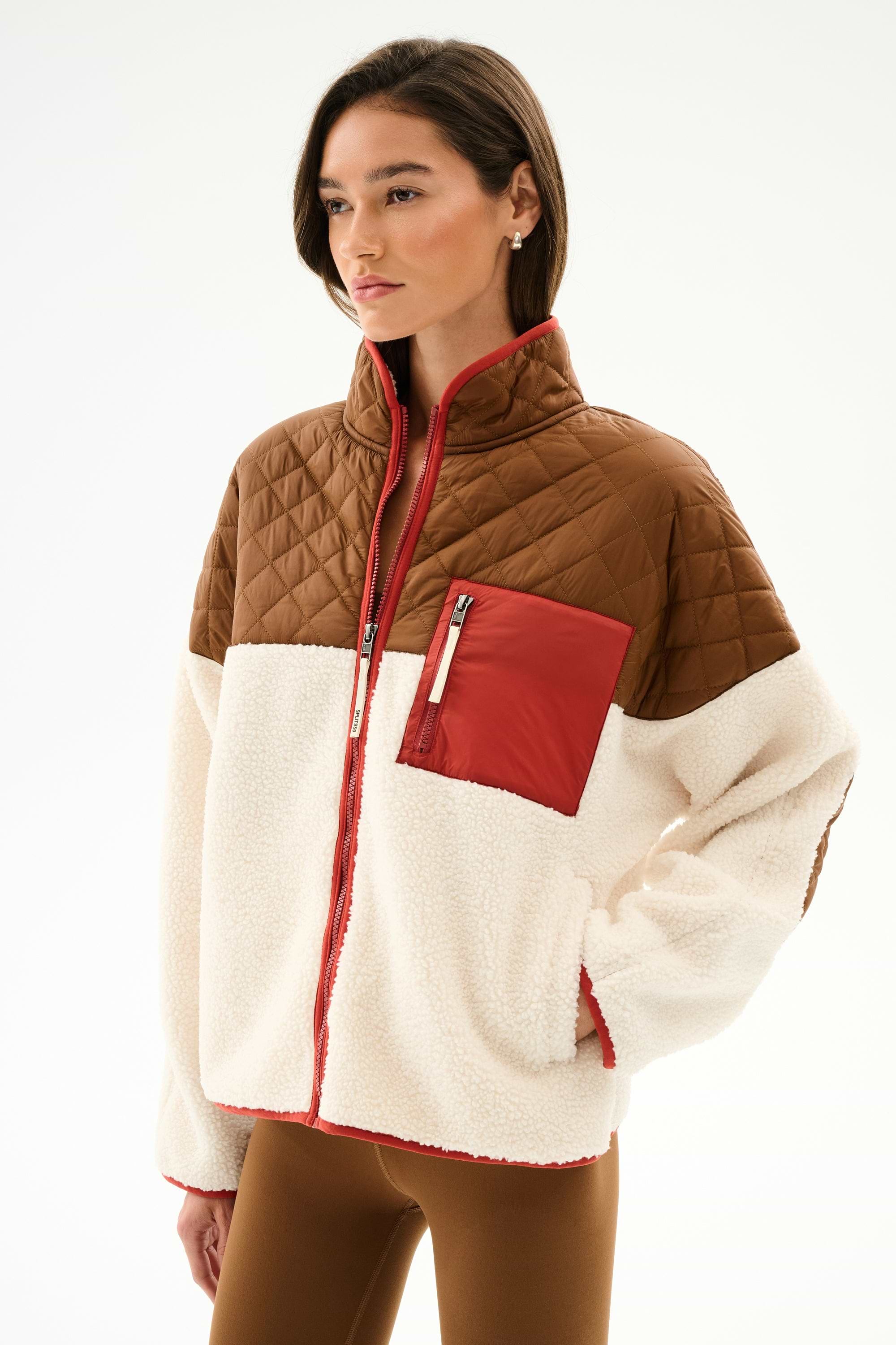 Woman in the SPLITS59 Skylar Sherpa Jacket - Creme/Toffee with brown quilted shoulders, red chest pocket, and cream fleece body, styled with brown pants.
