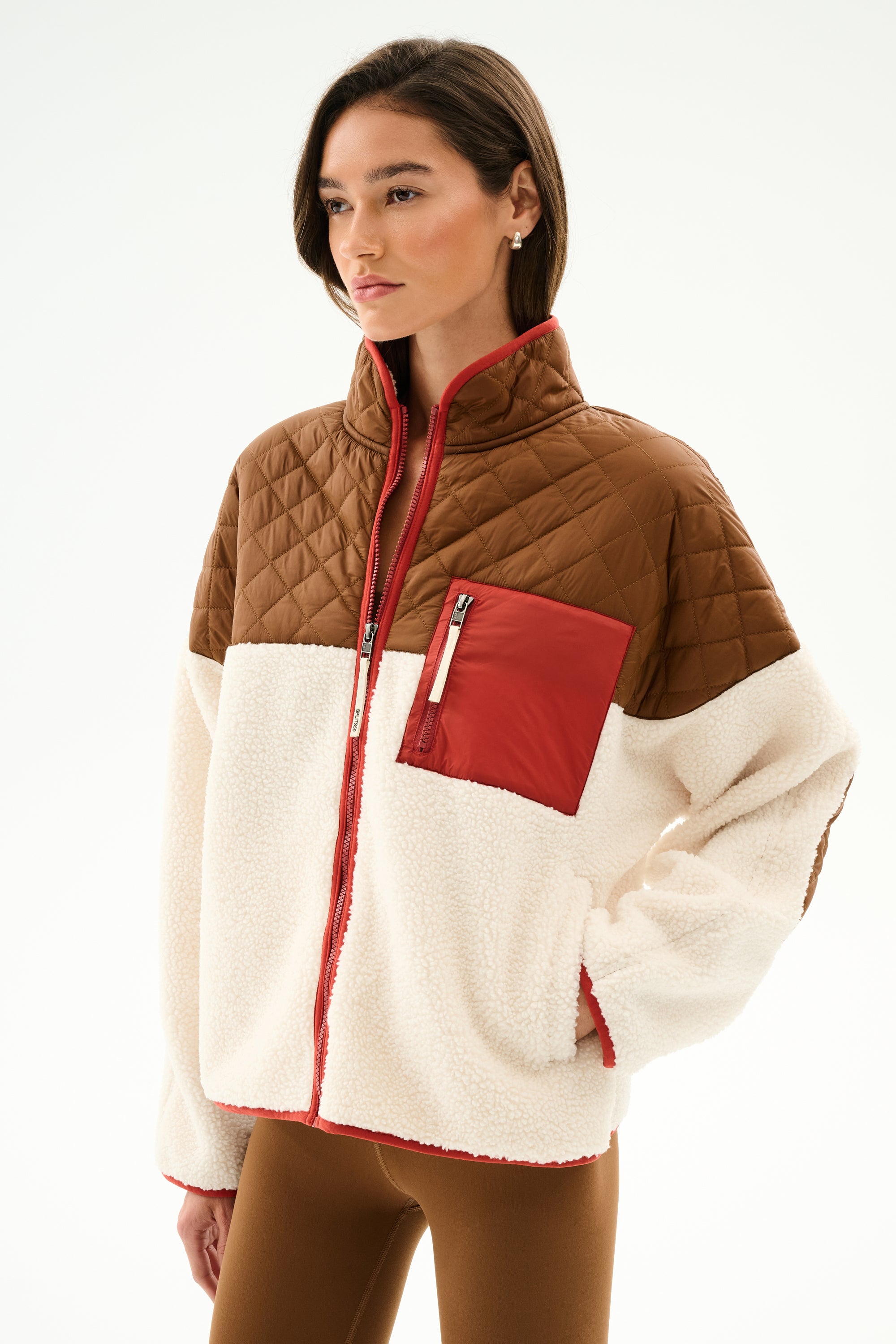 Woman in the SPLITS59 Skylar Sherpa Jacket - Creme/Toffee with brown quilted shoulders, red chest pocket, and cream fleece body, styled with brown pants.