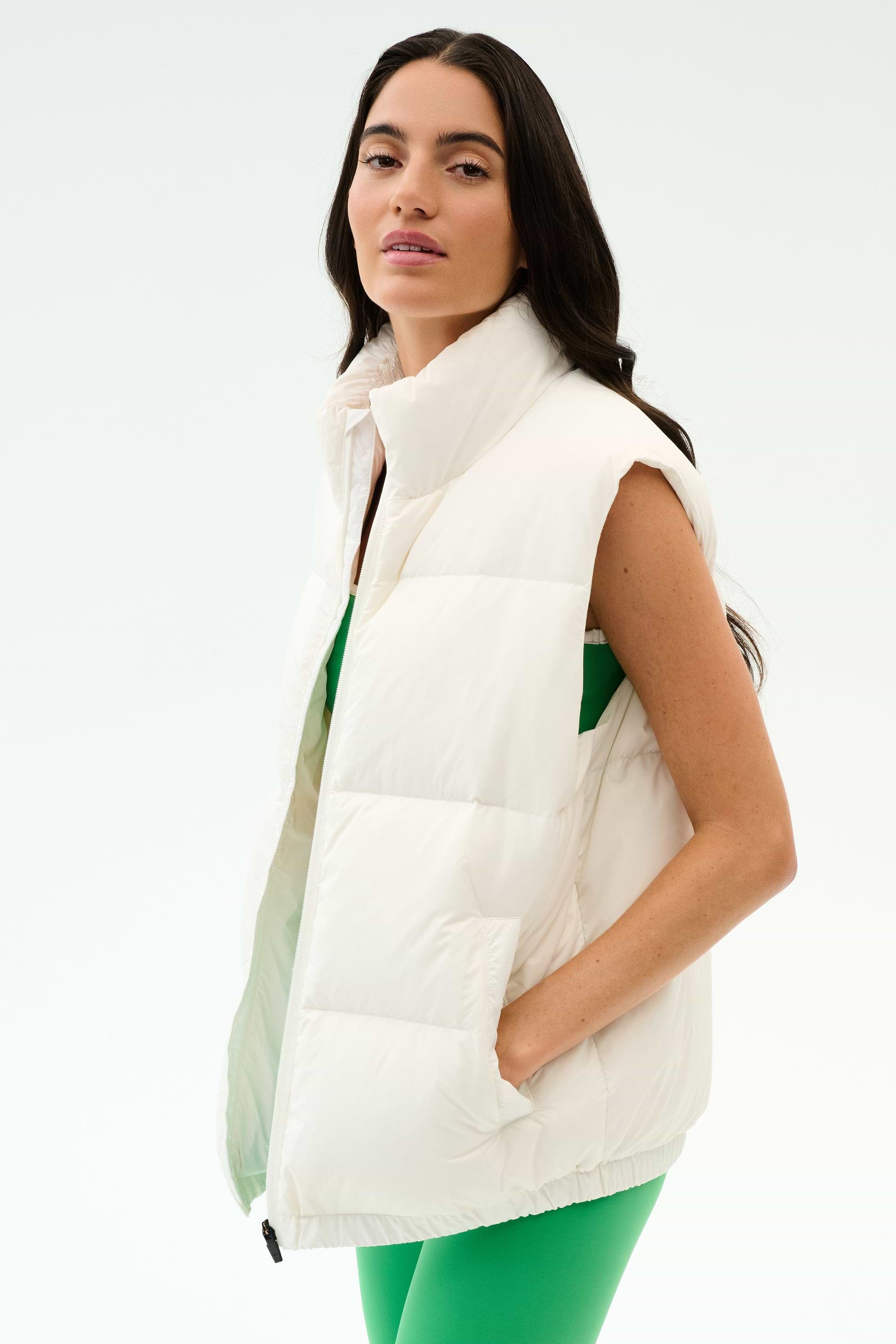 A woman sports the SPLITS59 Summit Nylon Puffer Vest in white over a green outfit, standing with one hand in her pocket against a plain backdrop—perfect lightweight insulation for cool weather workouts.