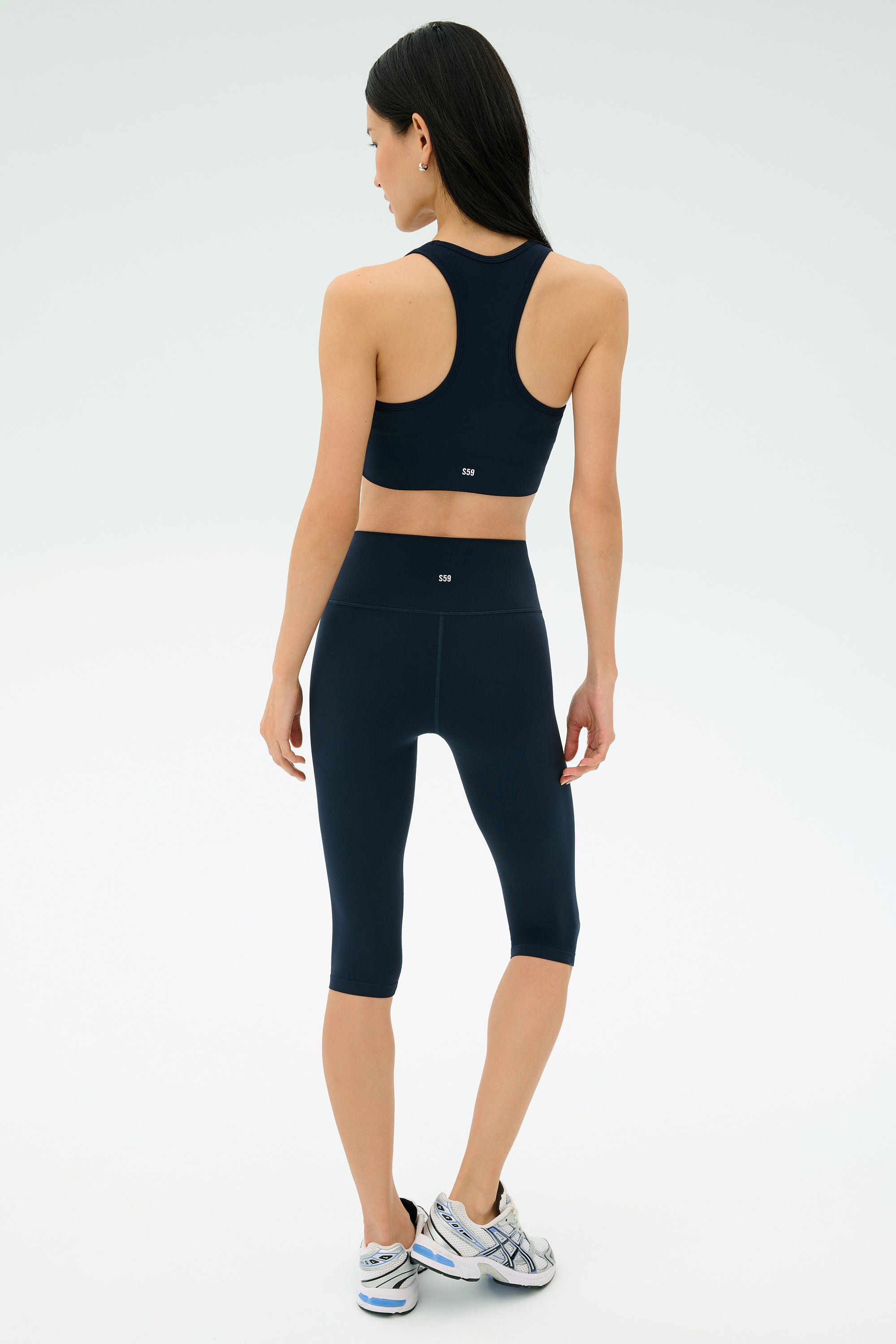 A woman stands facing away, wearing a navy SPLITS59 Airweight High Waist 16" Legging in Indigo and a sports bra with silver athletic shoes against a plain light background.