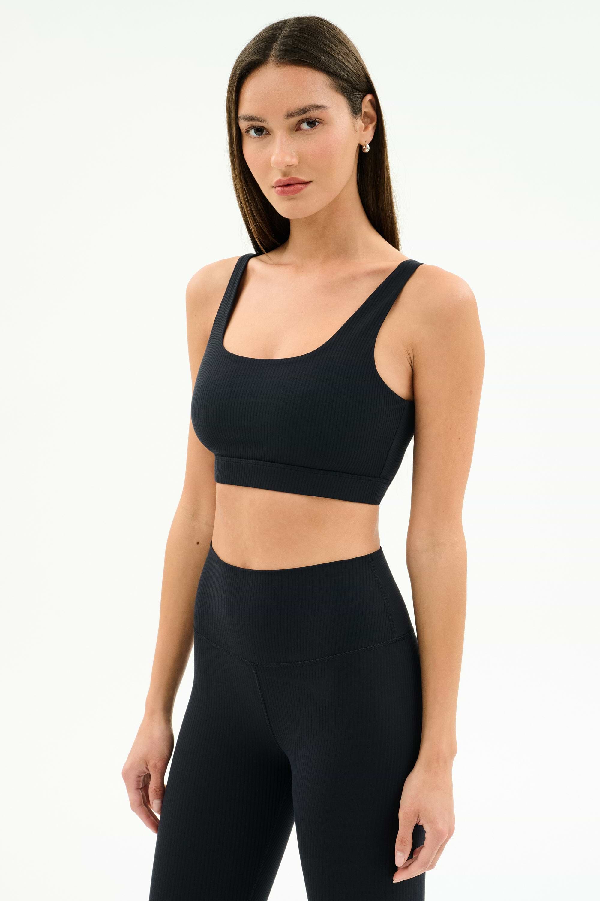 A woman with straight brown hair wears the SPLITS59 Airweight Rib Bra in black, paired with high-waisted leggings, both made from moisture-wicking fabric, standing against a plain white background.