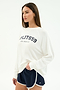 A woman wears a white SPLITS59 Andie Oversized Fleece Varsity Graphic Sweatshirt with "Splits59 Los Angeles" and navy athletic shorts, standing against a plain white background.