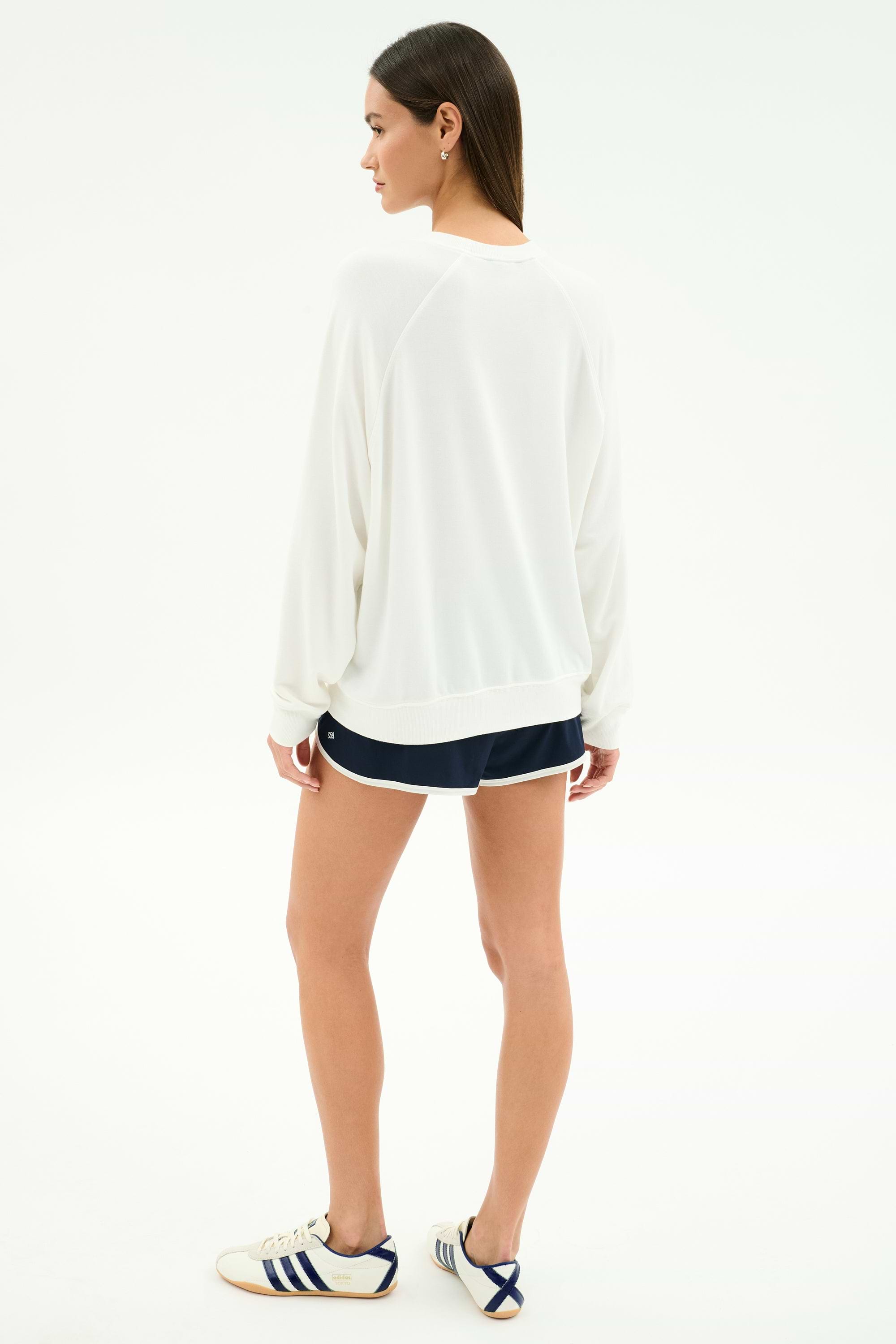 A person stands on a white background, facing away, wearing the SPLITS59 Andie Oversized Fleece Varsity Graphic Sweatshirt in white. This casually refined look is made in Los Angeles.