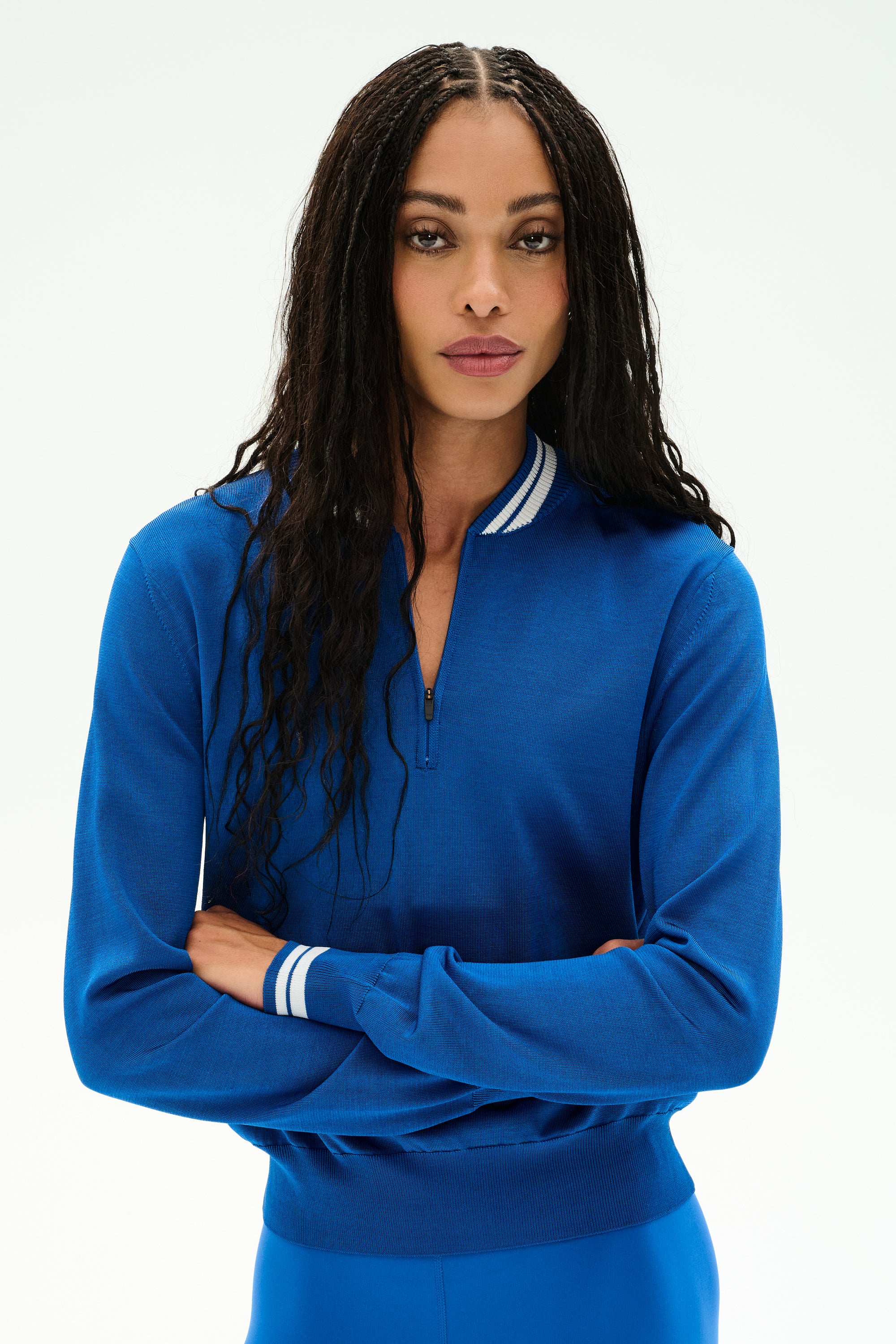 A person with long curly hair stands with arms crossed against a plain white background, wearing the SPLITS59 Bonnie Knit Half Zip in Classic Blue/White—a bright blue sweater crafted from a soft rayon and nylon blend, detailed with white stripes on the collar and cuffs.